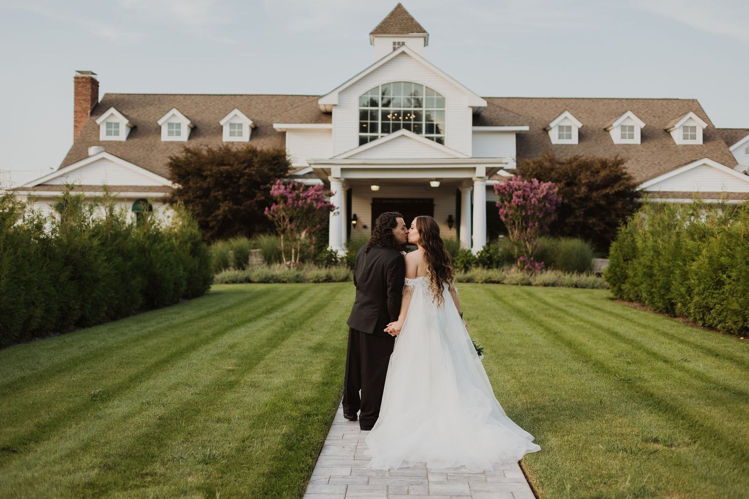 A couple in formal wedding attire walking away on a manicured lawn path leading to an elegant white mansion with dormers.