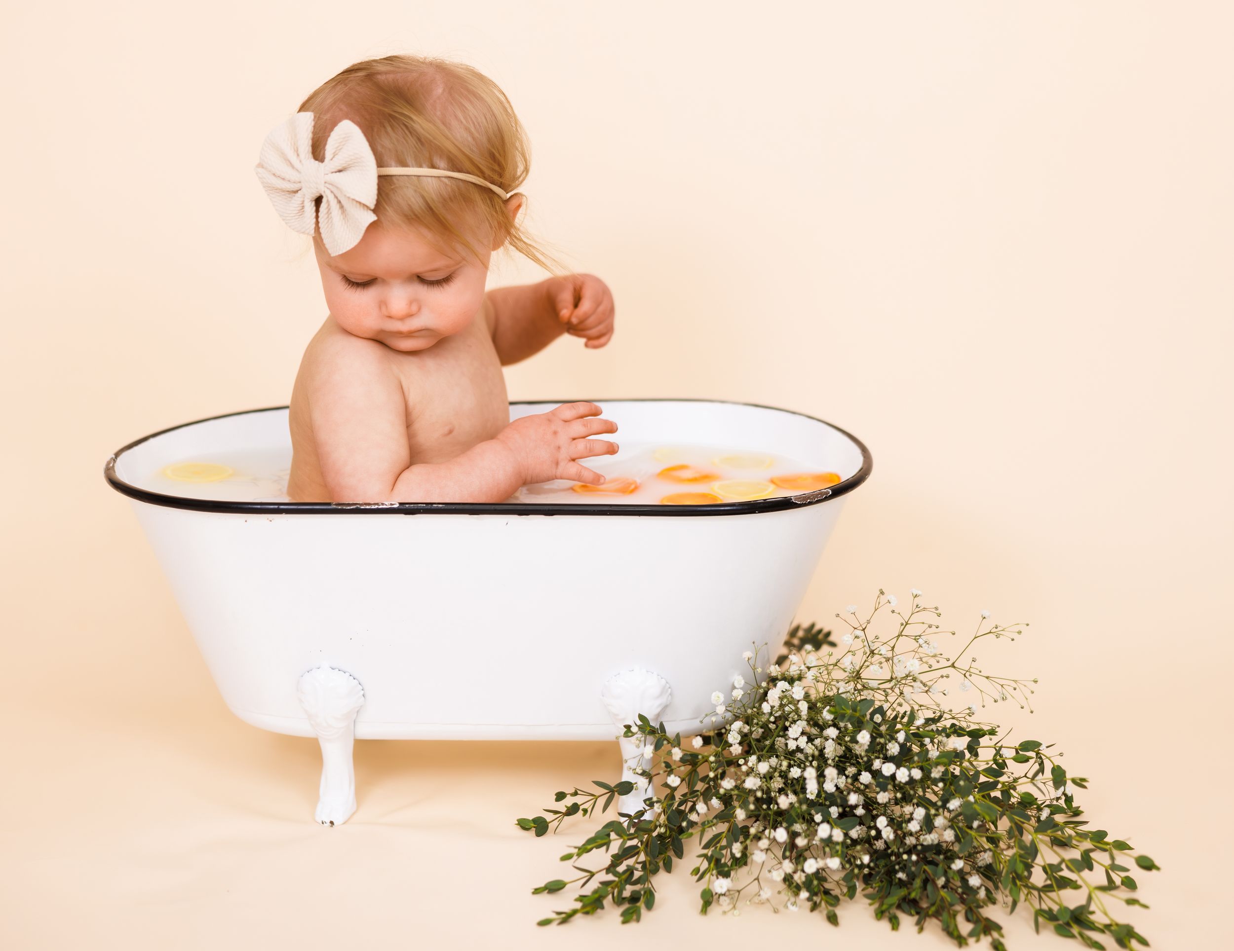 A baby sits in a vintage white enamel bathtub with flowers beside it against a peachy background.