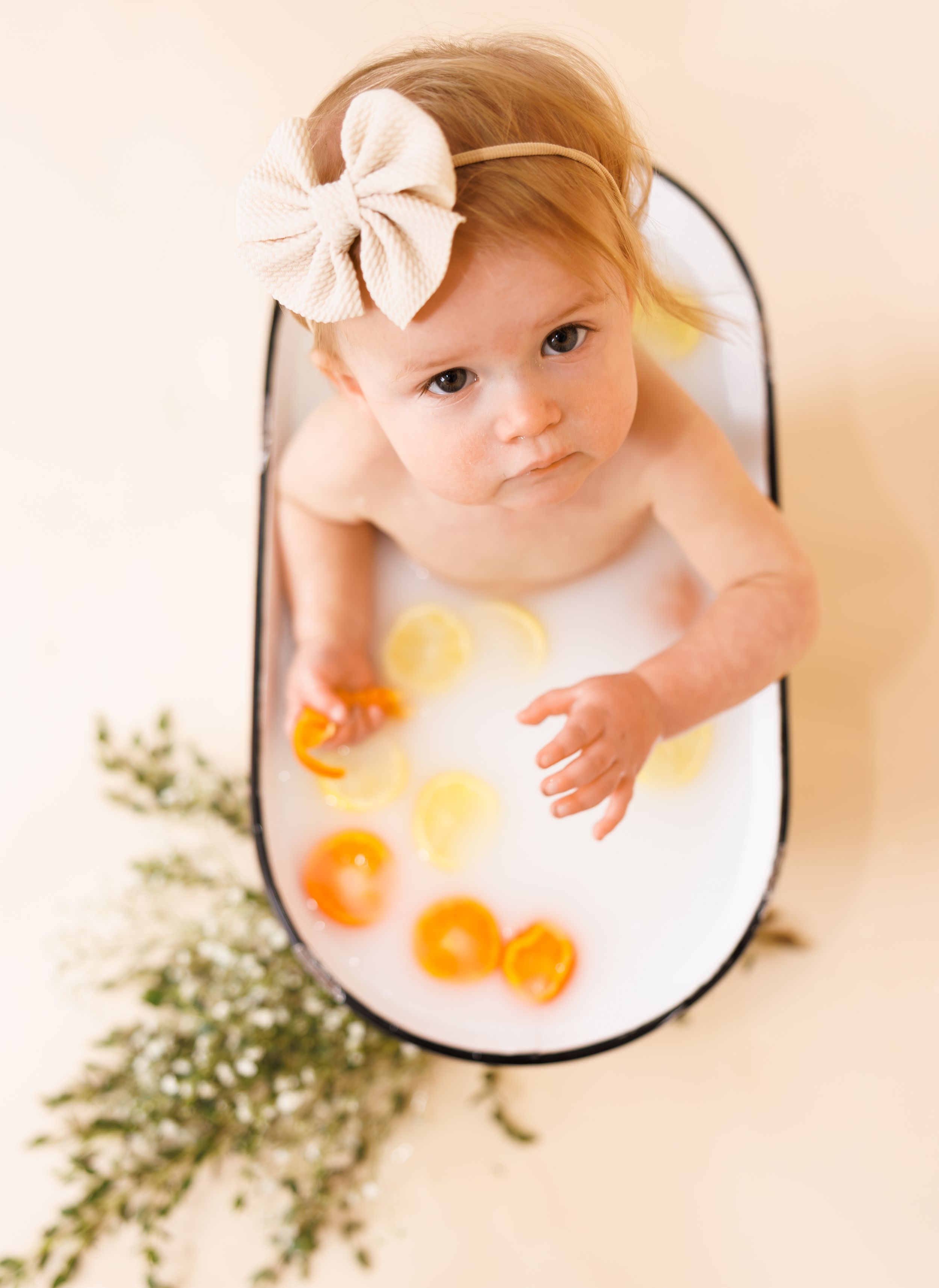 Overhead milk bath photography with orange citrus fruits and greenery against a peachy background and white fabric.