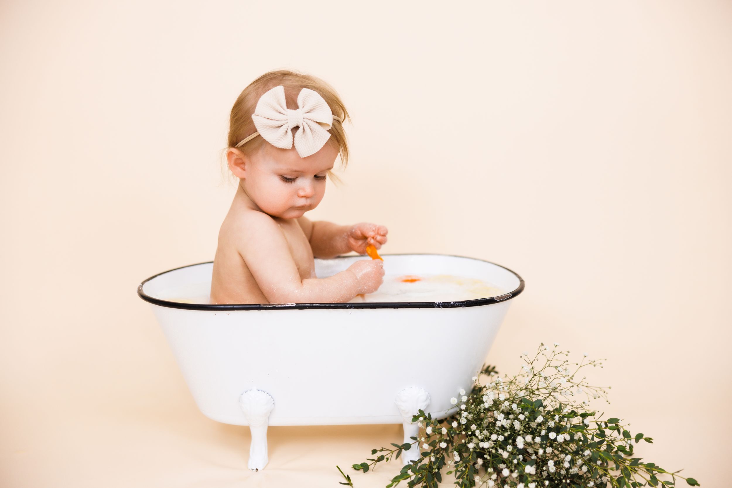 A baby wearing a white bow sits in a miniature white bathtub next to fresh baby's breath flowers against a peach backdrop.