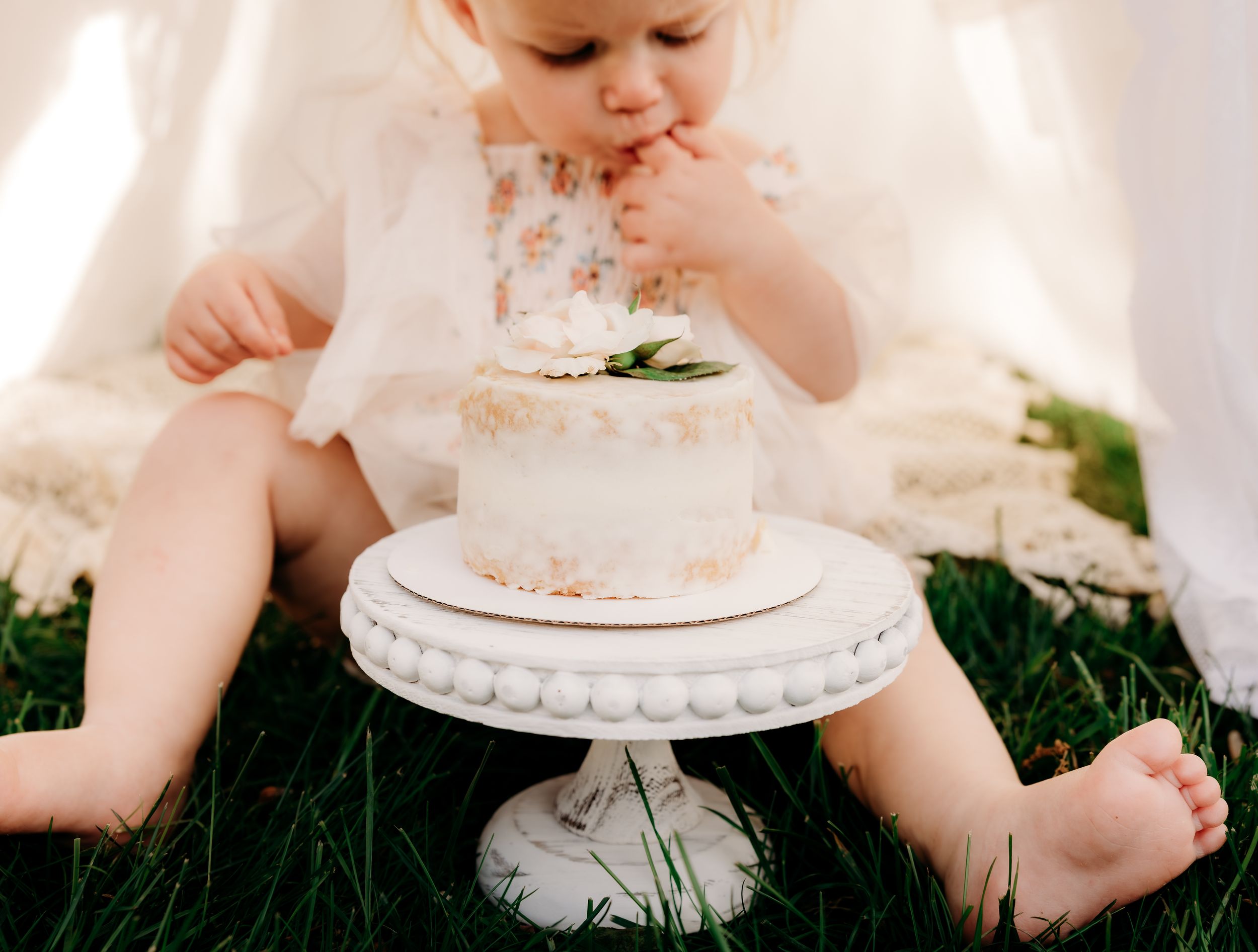 Small child sitting on grass tasting frosting from a white cake displayed on an ornate pedestal stand.