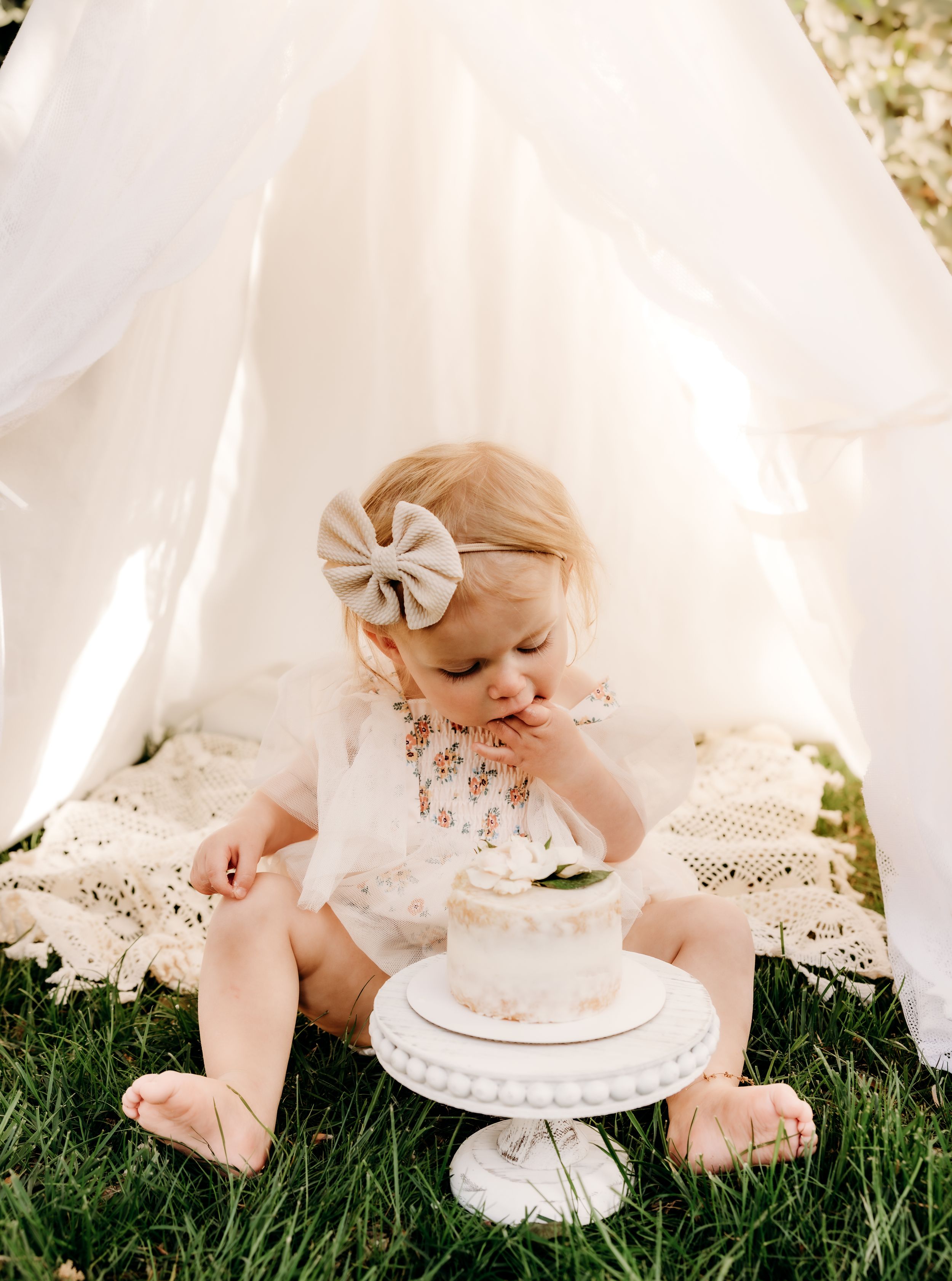 A small child in white lace sits on grass with a white cake under a sheer canopy tent.