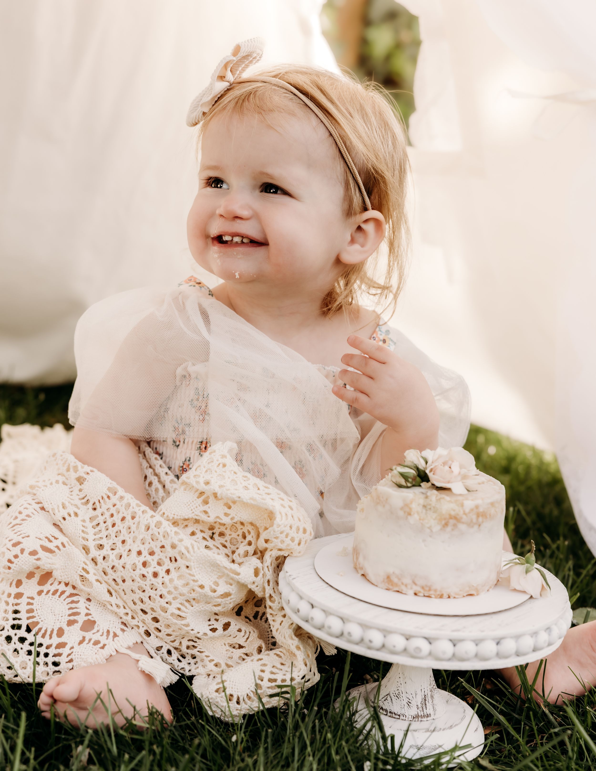 Happy toddler in white lace dress sits on grass beside small decorated white cake on ornate pedestal stand.