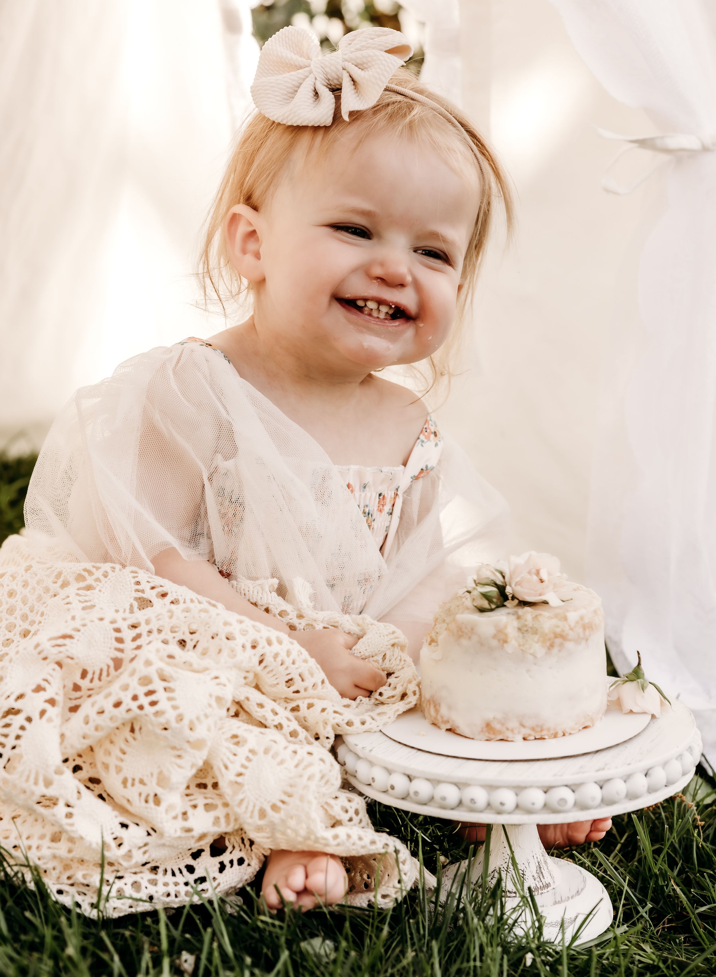 Baby in lace dress sitting on grass with delicate white cake on decorative pedestal stand.