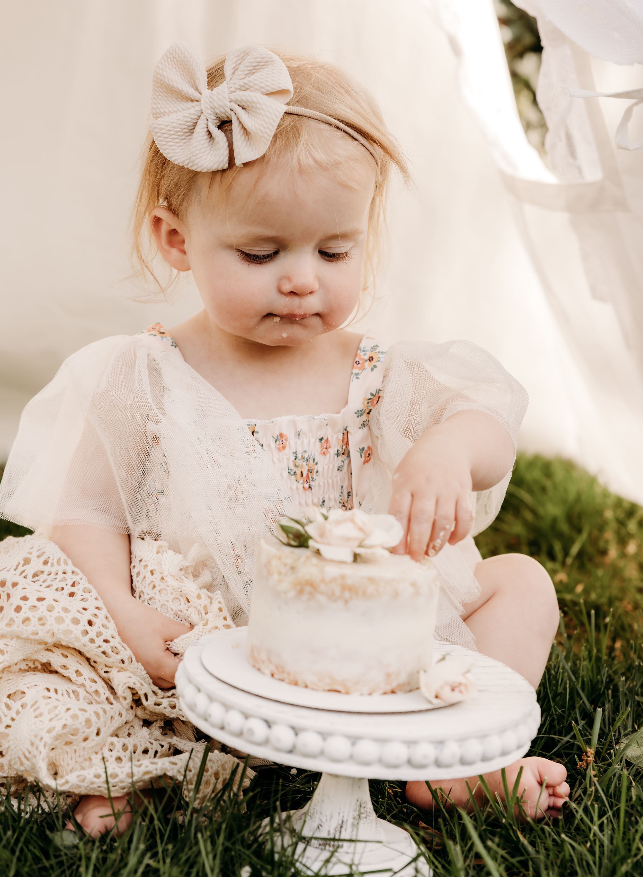 A small child in a cream lace dress sits on grass reaching for a delicate white cake displayed on an elegant pedestal stand.