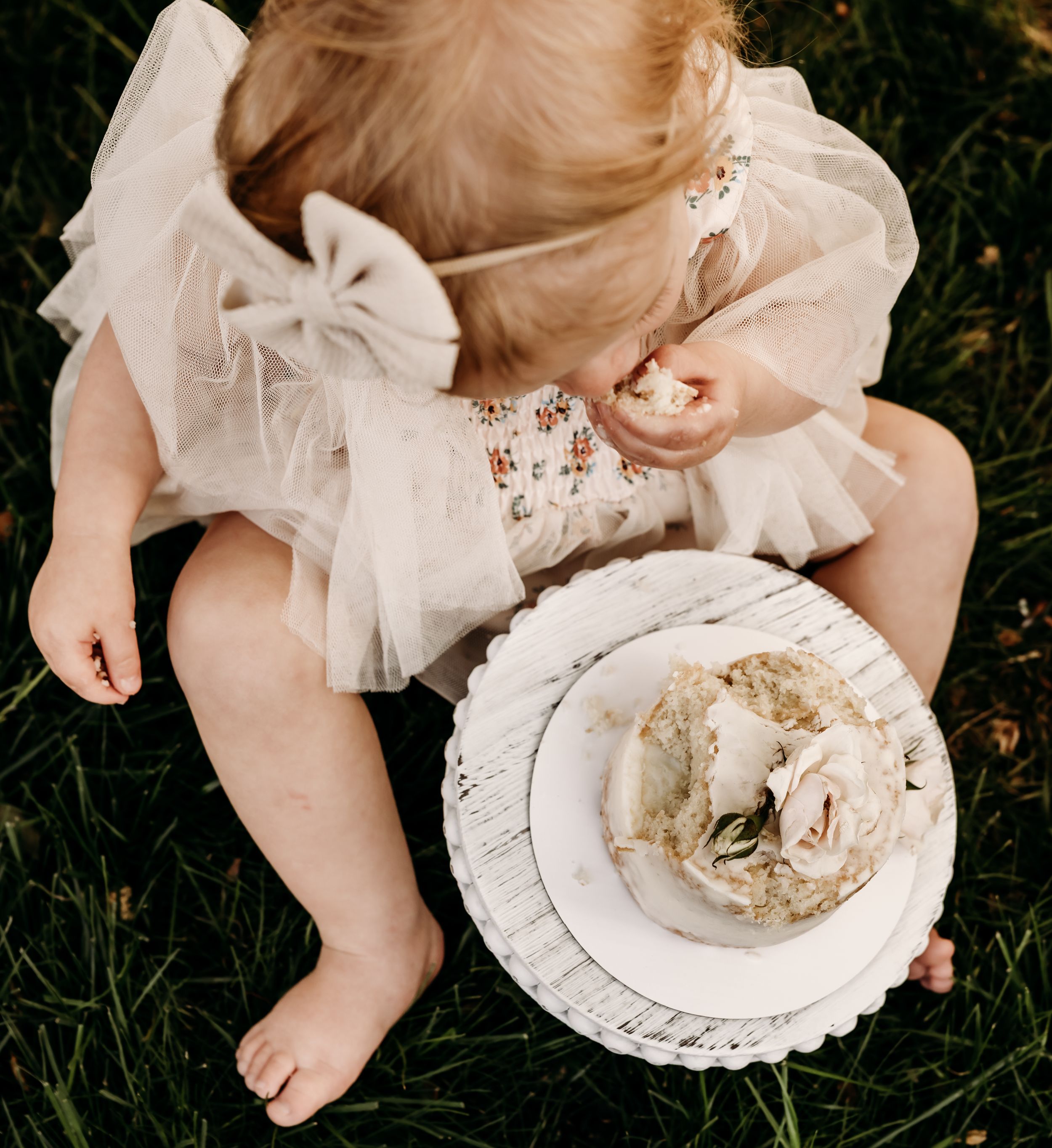 High angle view of small child in white dress sitting on grass next to vintage cake plate with floral decor.