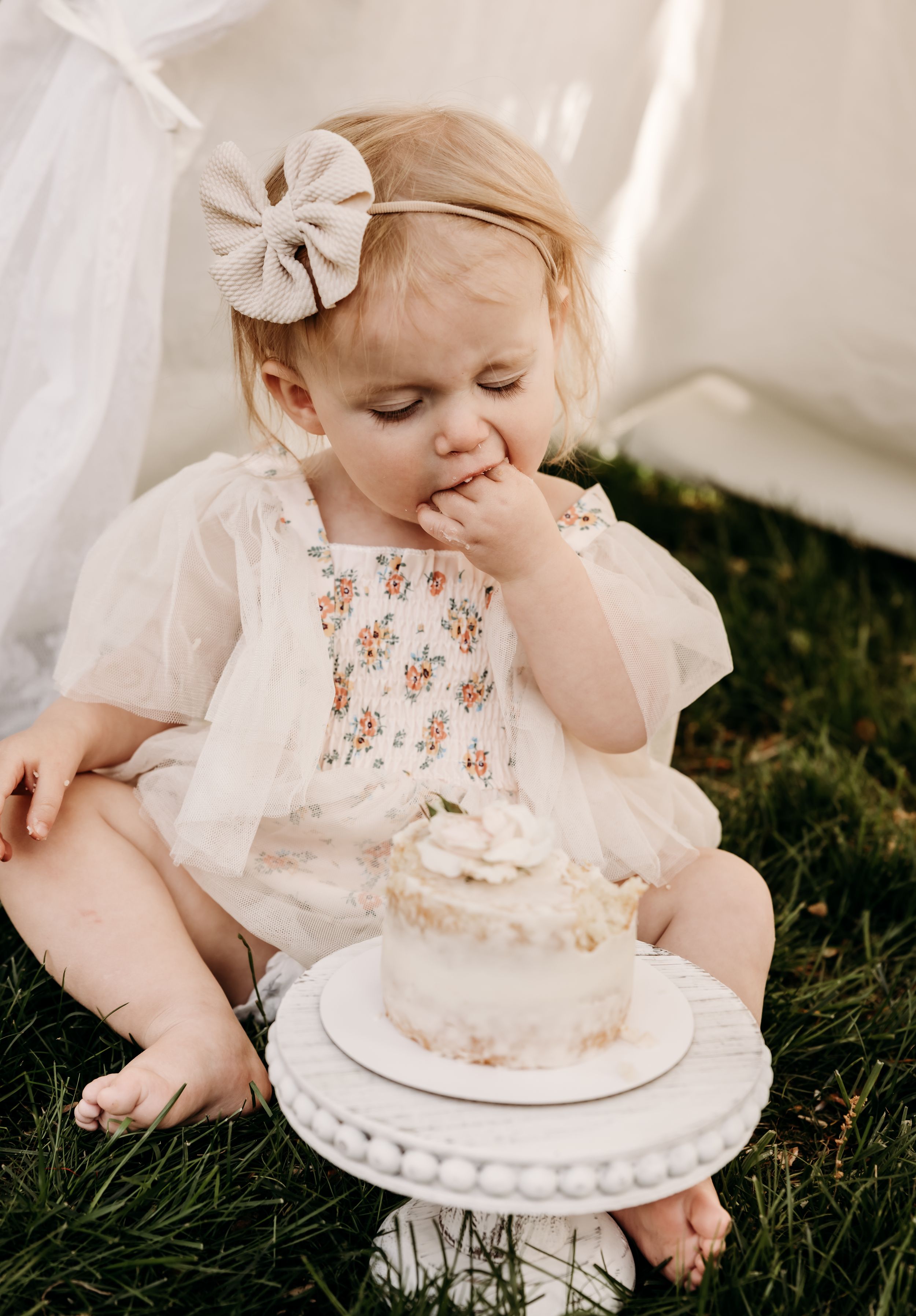 Sweet baby in white dress sits on grass with cake during first birthday cake smash photoshoot with vintage styling.