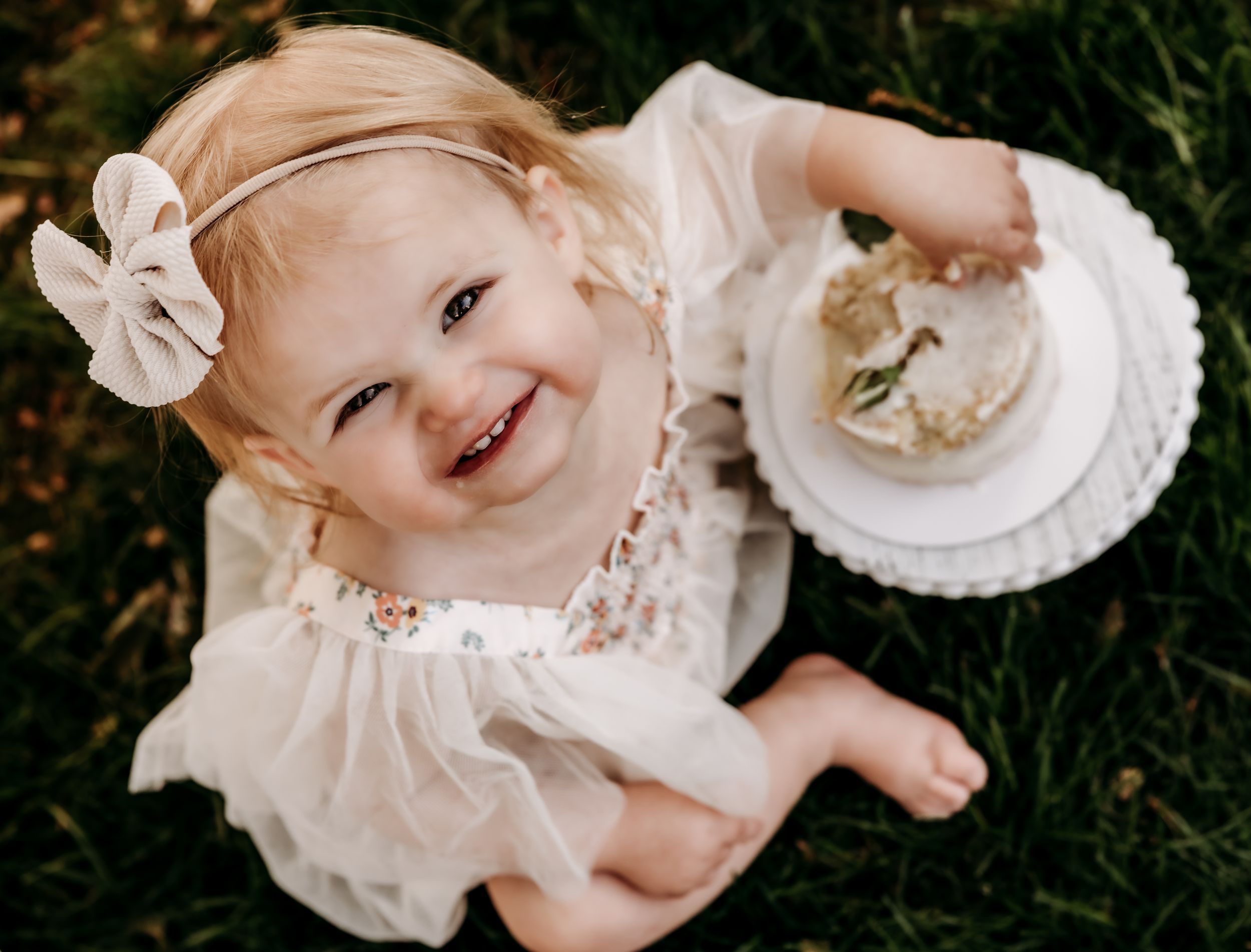 A toddler in a white dress sits on grass while smiling and holding a plate.