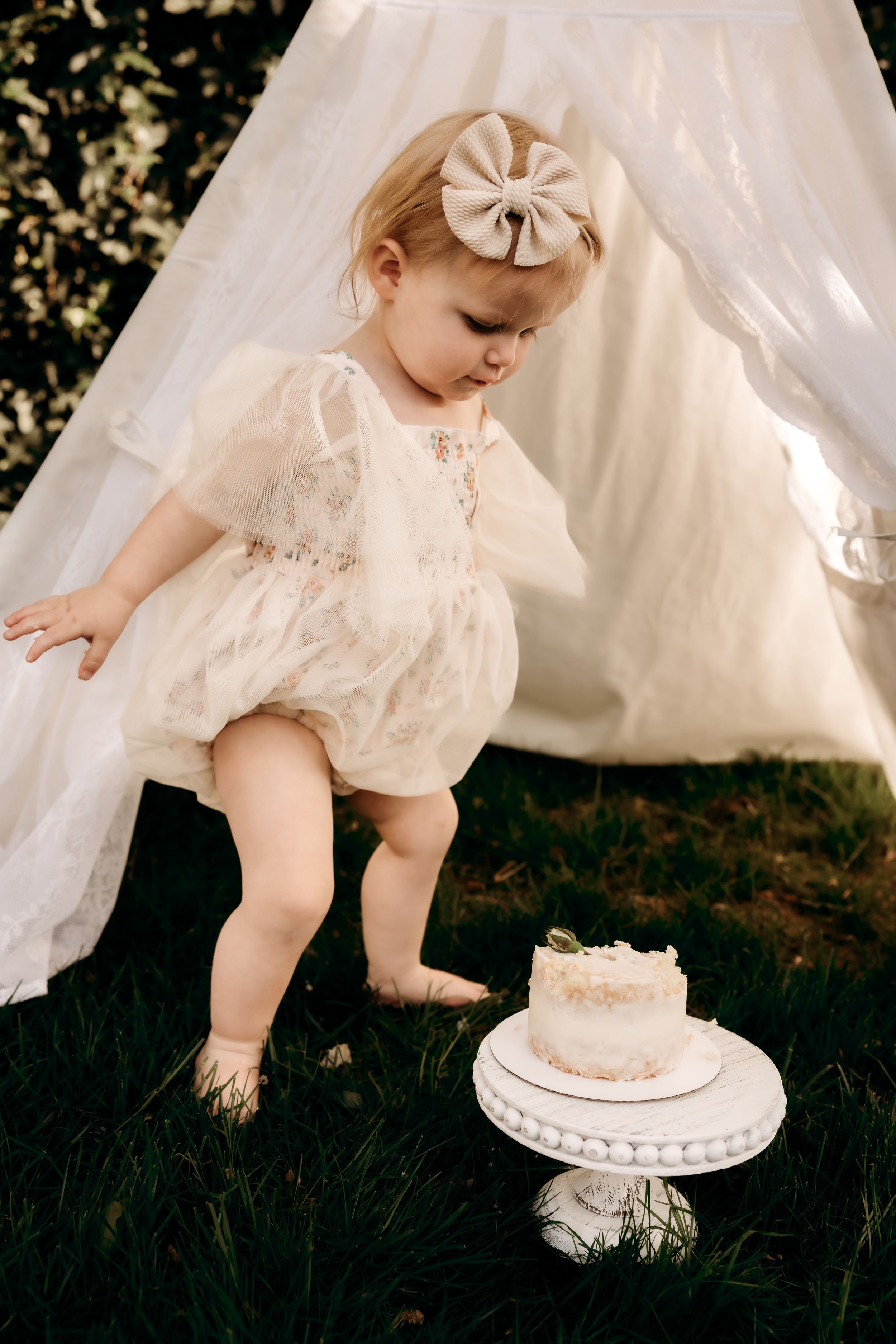 A baby in a cream lace dress reaches toward a small white cake beside a white tent setup in a garden setting.