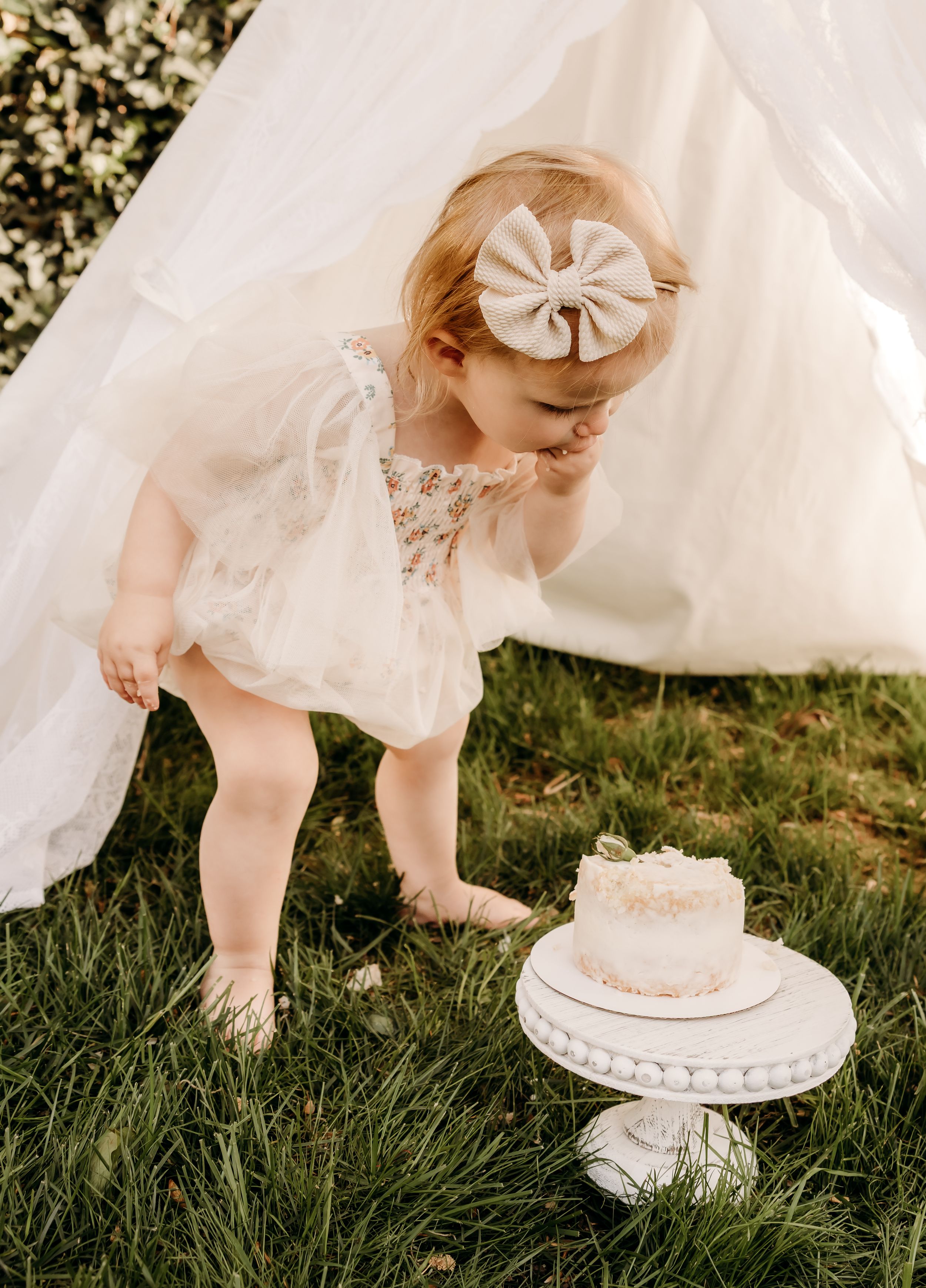 A small baby in a lacy white dress and bow reaches down towards a delicate white cake on a pedestal stand in the grass.