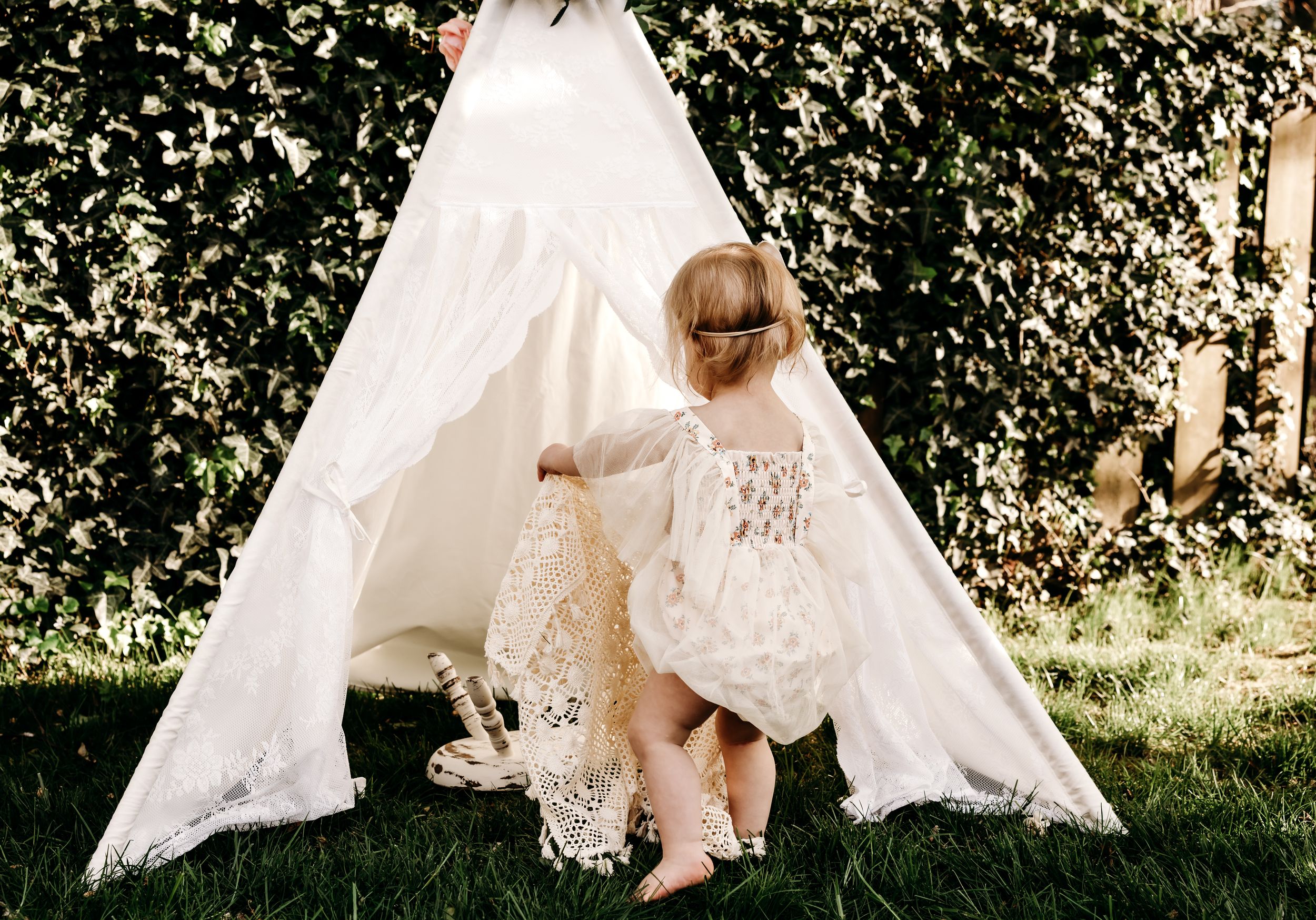 A white lace teepee tent set up on grass with green ivy covered wall in the background.