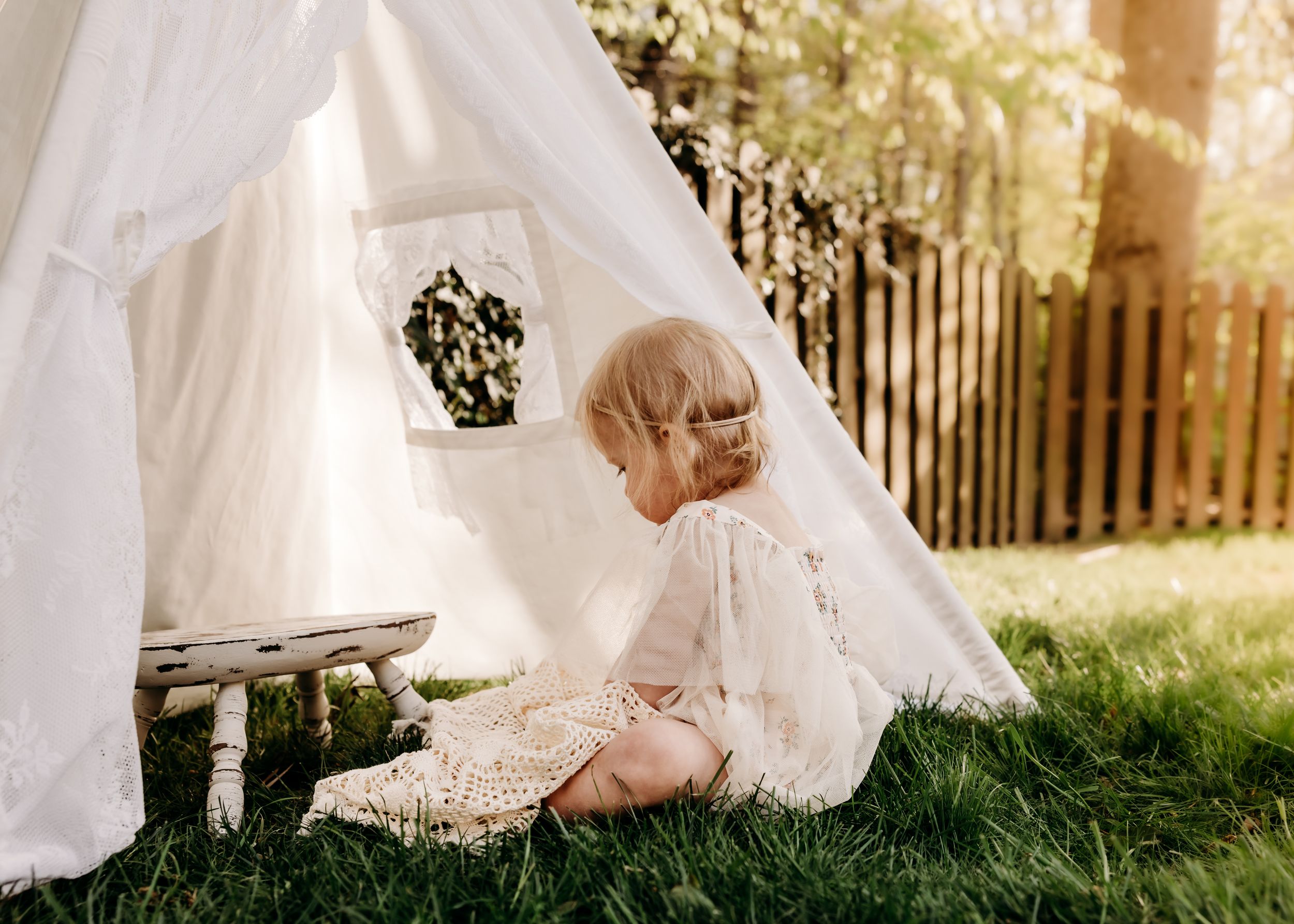 A small child in a white dress sits by a teepee tent in a sunlit backyard garden setting.