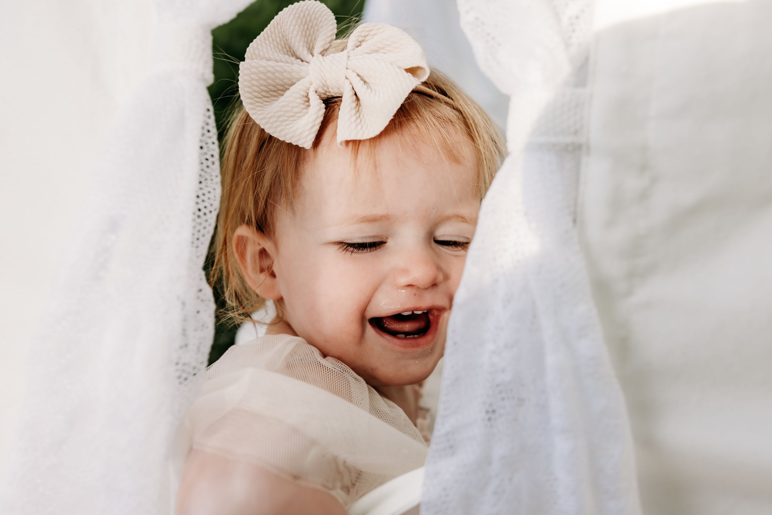 A smiling child wearing a cream colored bow and dress peeks through white fabric.