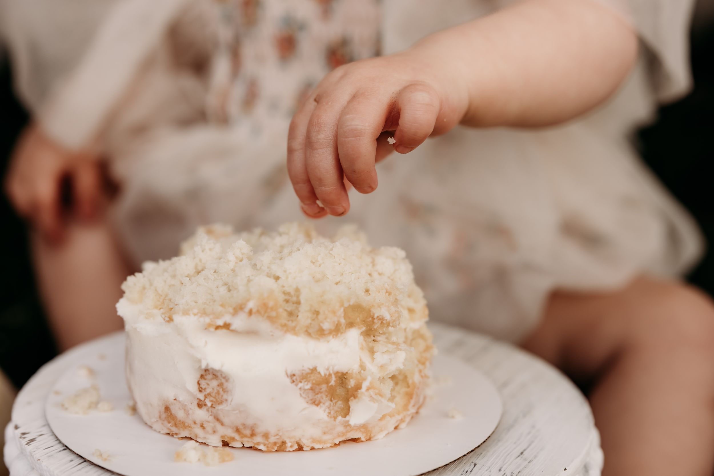 A small hand reaches for a white frosted cake decorated with coconut flakes on a white plate.