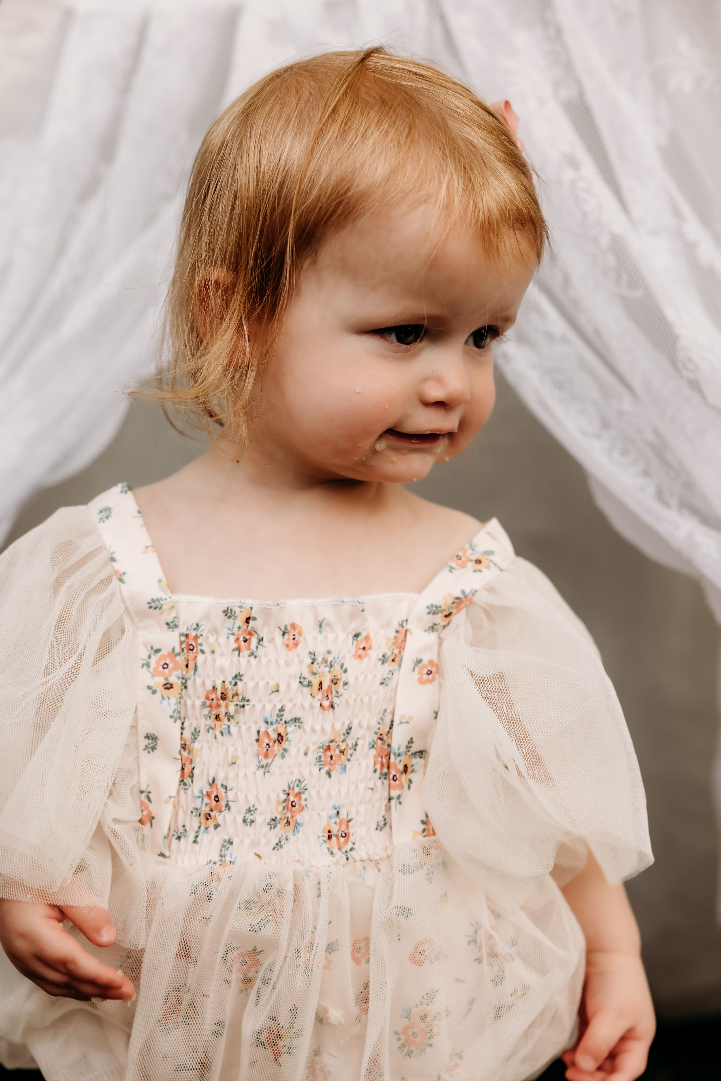 A toddler wearing a delicate cream dress with floral embroidery smiles gently against a white fabric backdrop.