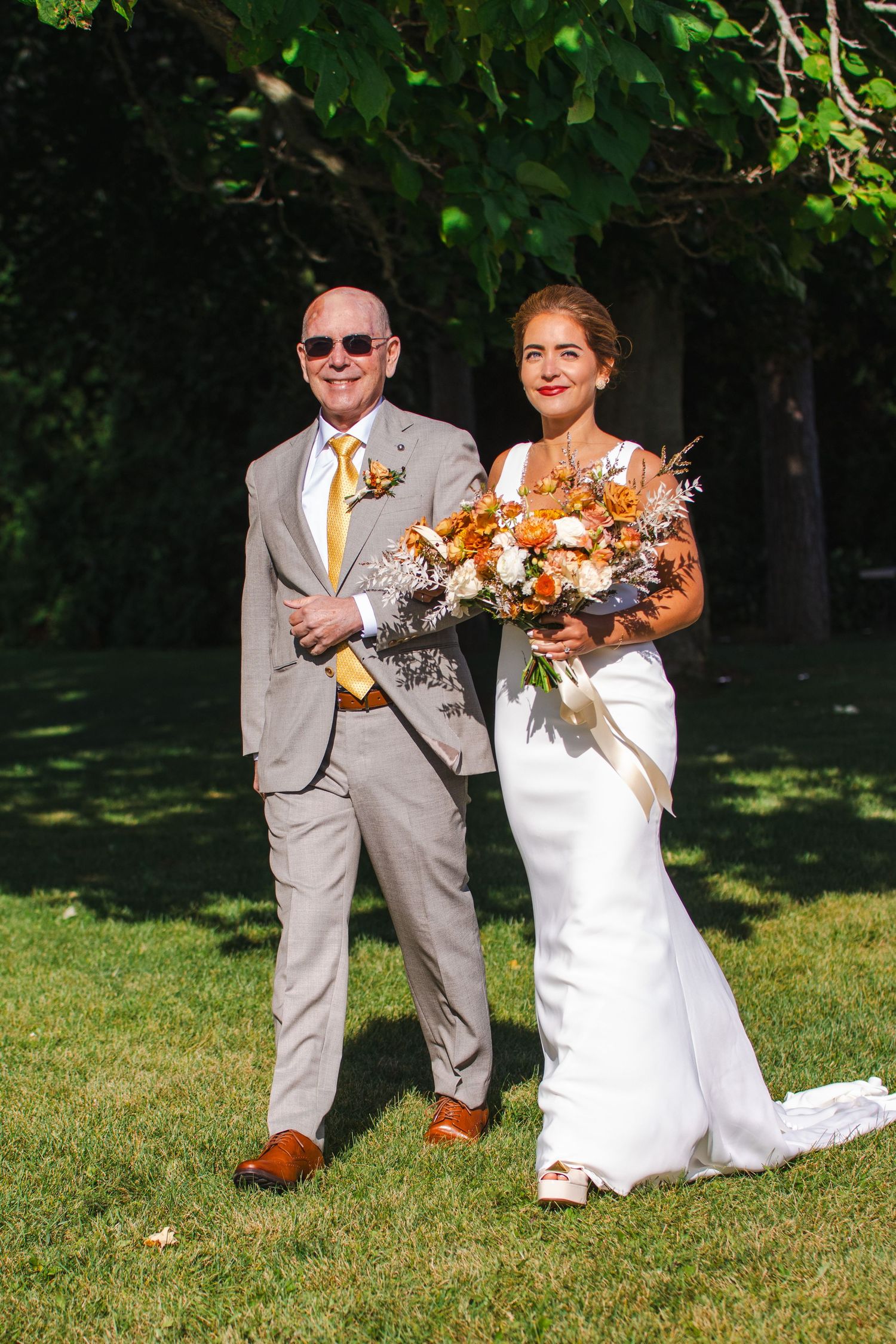 Wedding couple stands on lawn during summer ceremony with bride in white dress holding orange and white bouquet.