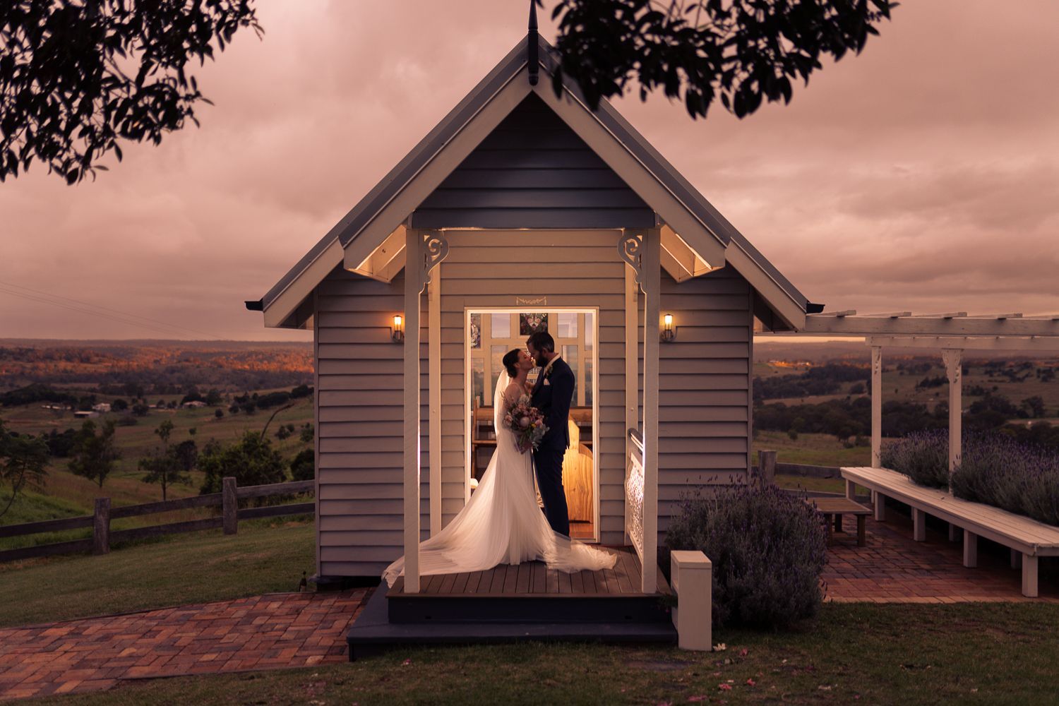 A couple is standing in the doorway of a small, charming chapel at sunset. The scene is romantic, with a scenic landscape in the background