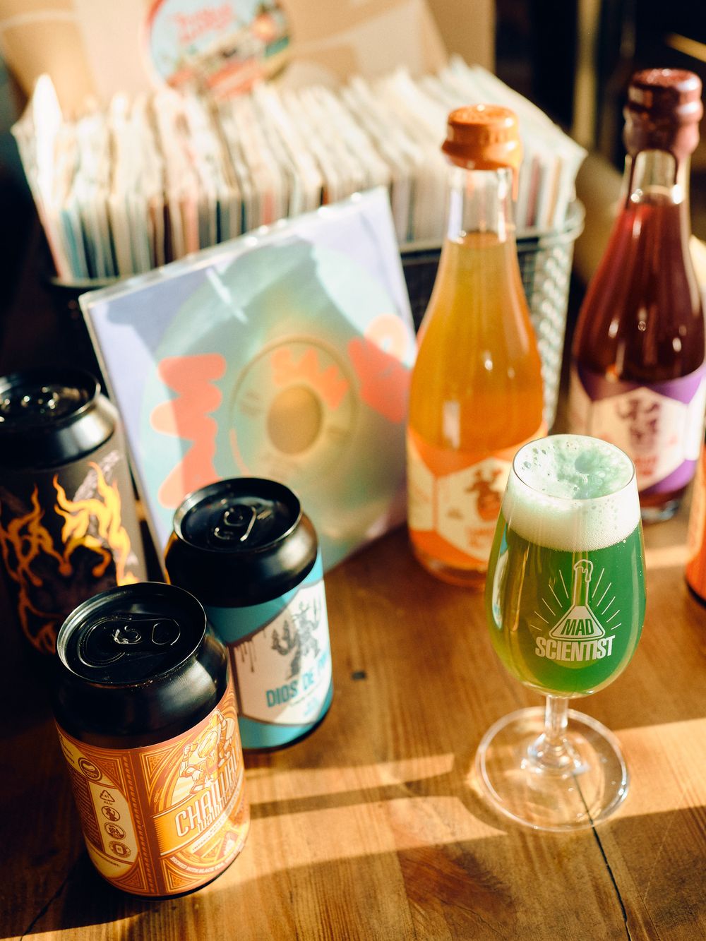 Craft beer and beverage display with colorful cans, glass bottles, and a filled beer glass on wooden surface.