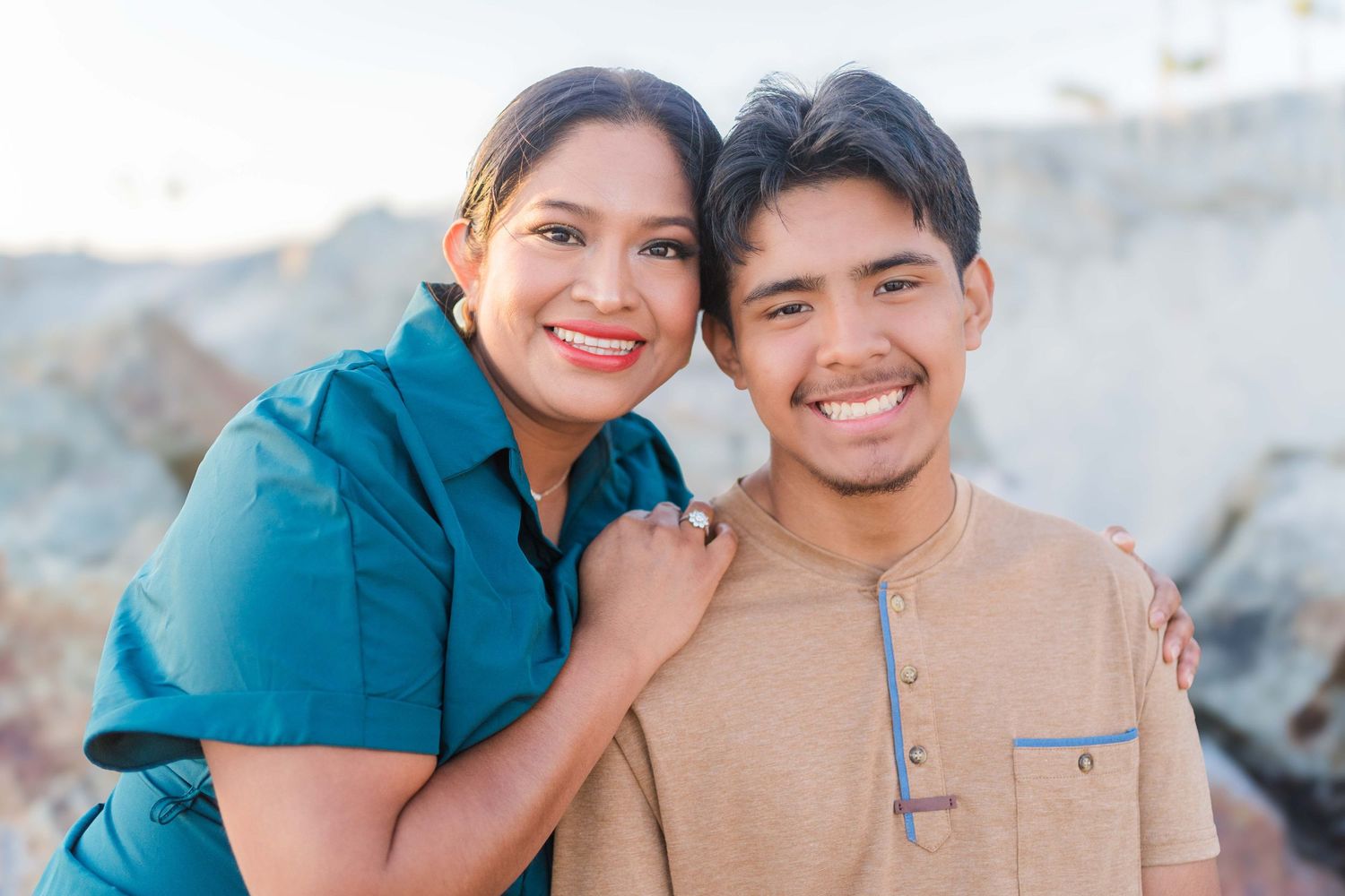 A couple embraces on the beach with rocky coastal formations in the background.