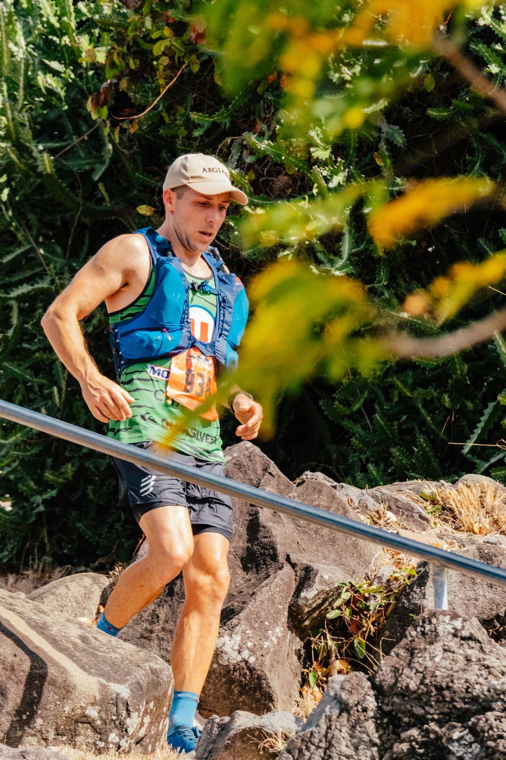 Trail runner in blue tank top and shorts navigates rocky terrain while carrying hydration gear during outdoor race.