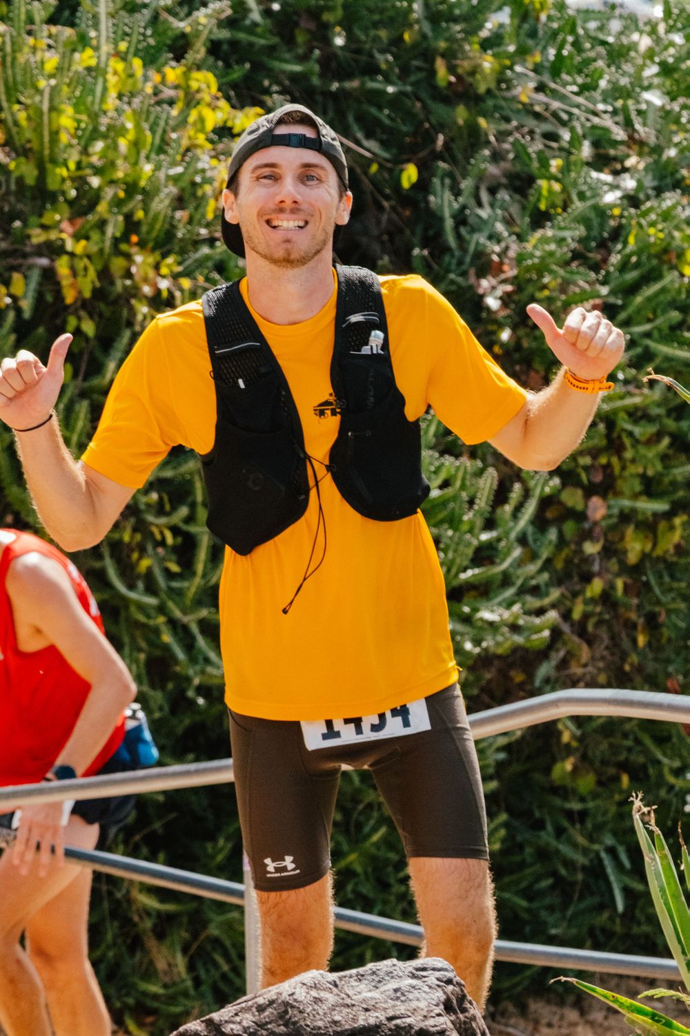 Athletic participant wearing yellow t-shirt and black vest making shaka gesture during outdoor trail race competition.
