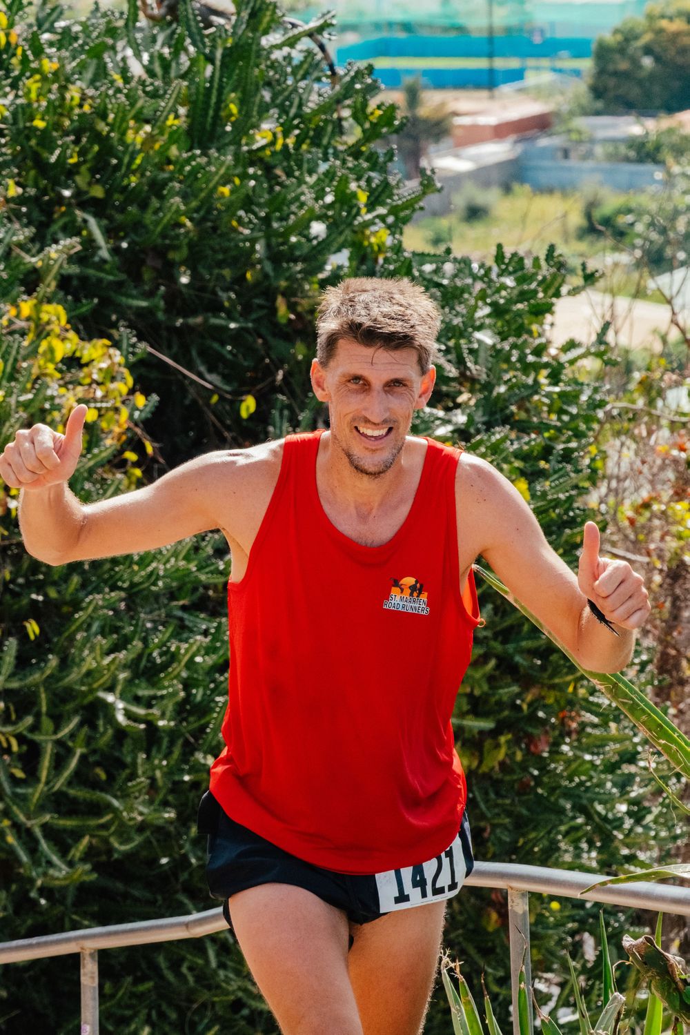 Athletic runner in a red tank top gives two thumbs up while standing on an outdoor path with greenery and ocean views.