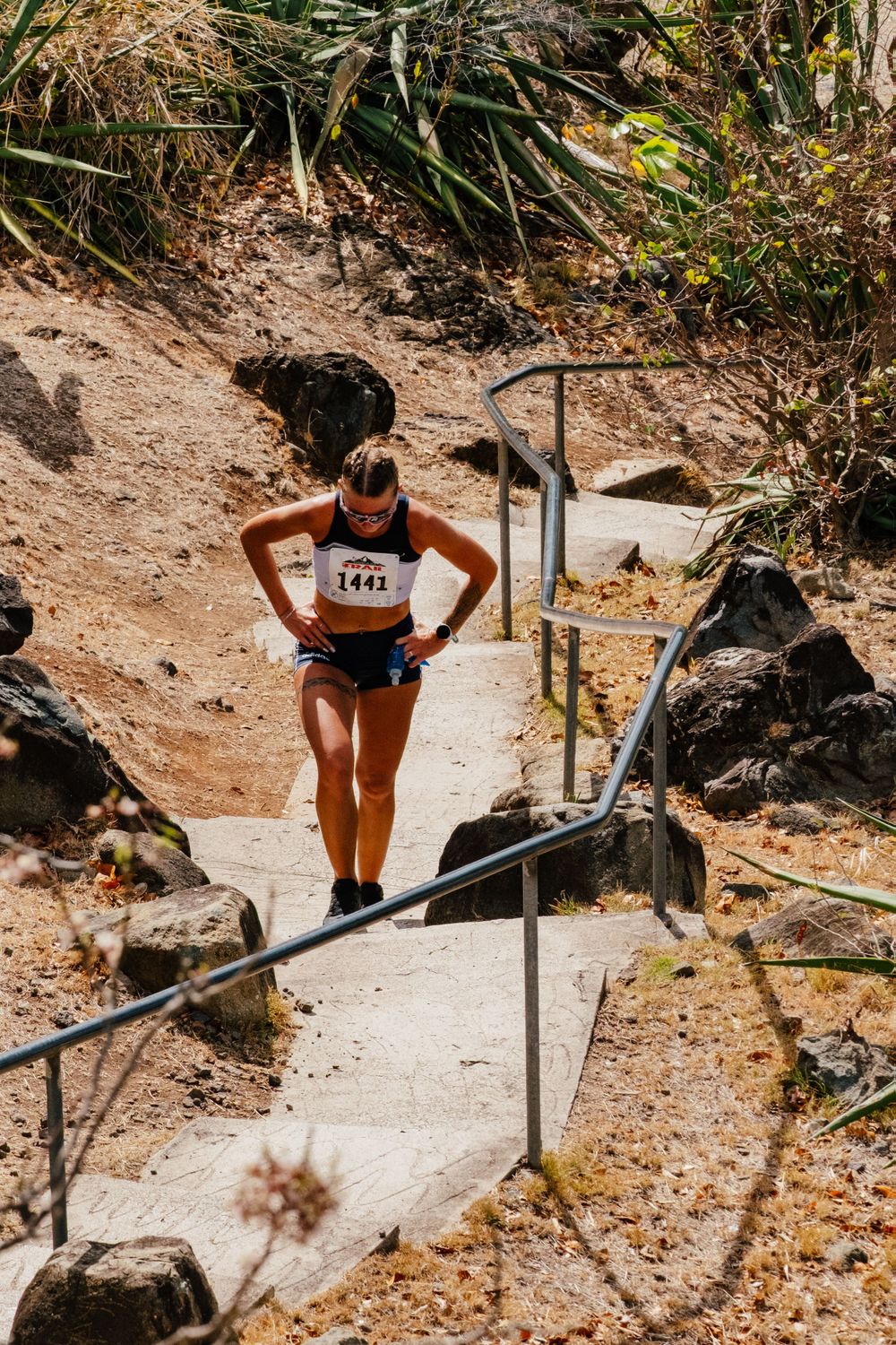 Runner on a rocky trail path with metal handrails pauses during an outdoor race in natural terrain.