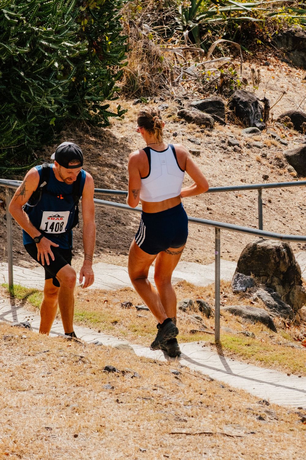 Two runners pause on a dirt trail with handrails during an outdoor trail running competition on a sunny day.
