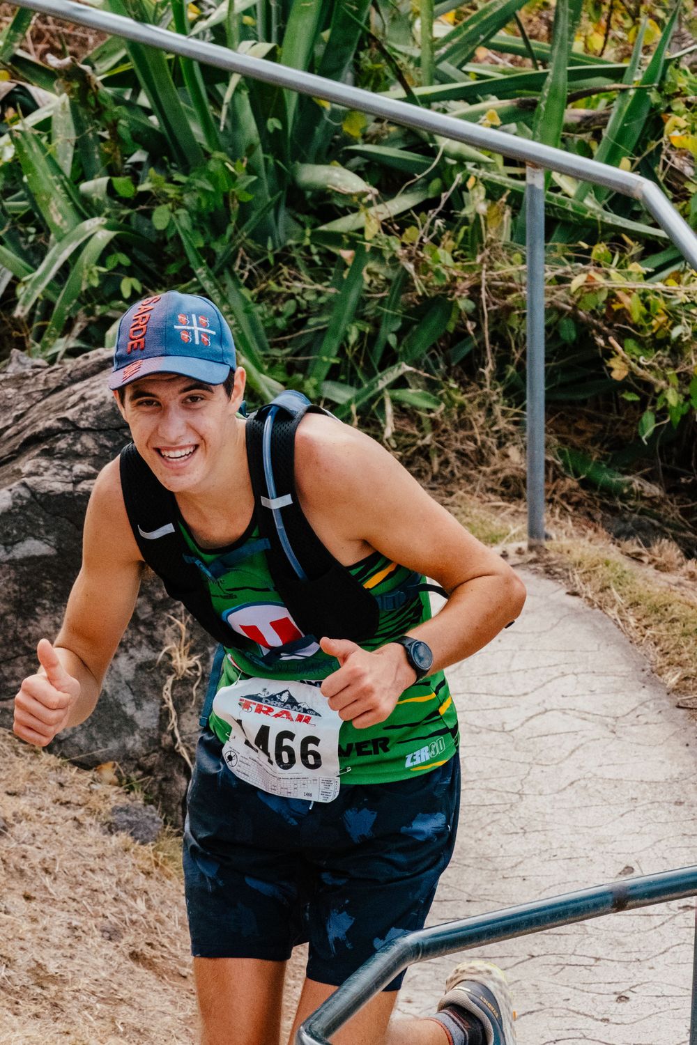 Trail runner wearing race number 466 gives thumbs up while ascending winding path surrounded by green foliage.