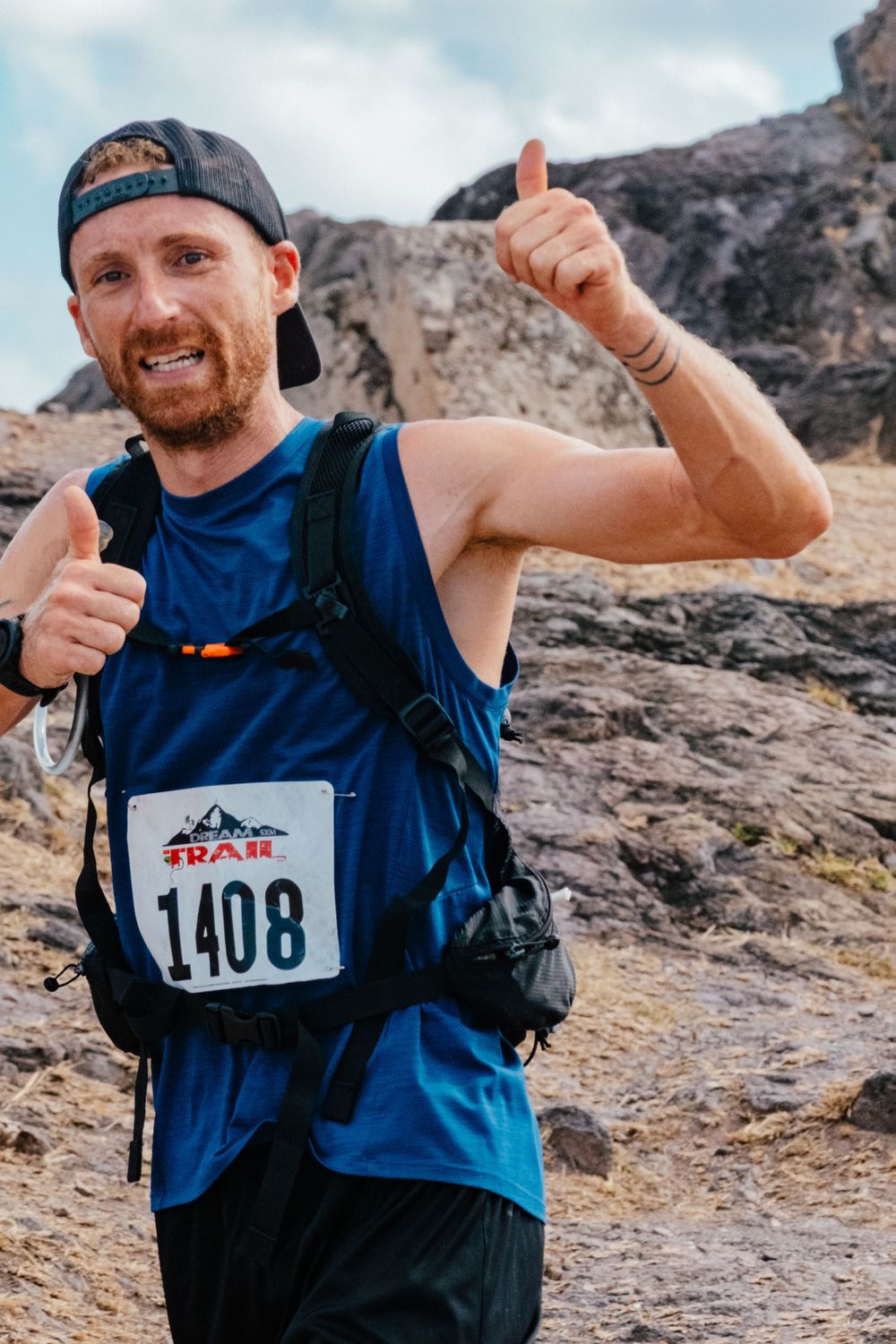 Trail runner wearing bib number 1408 gives thumbs up while hiking on rocky mountain terrain with a hydration pack.