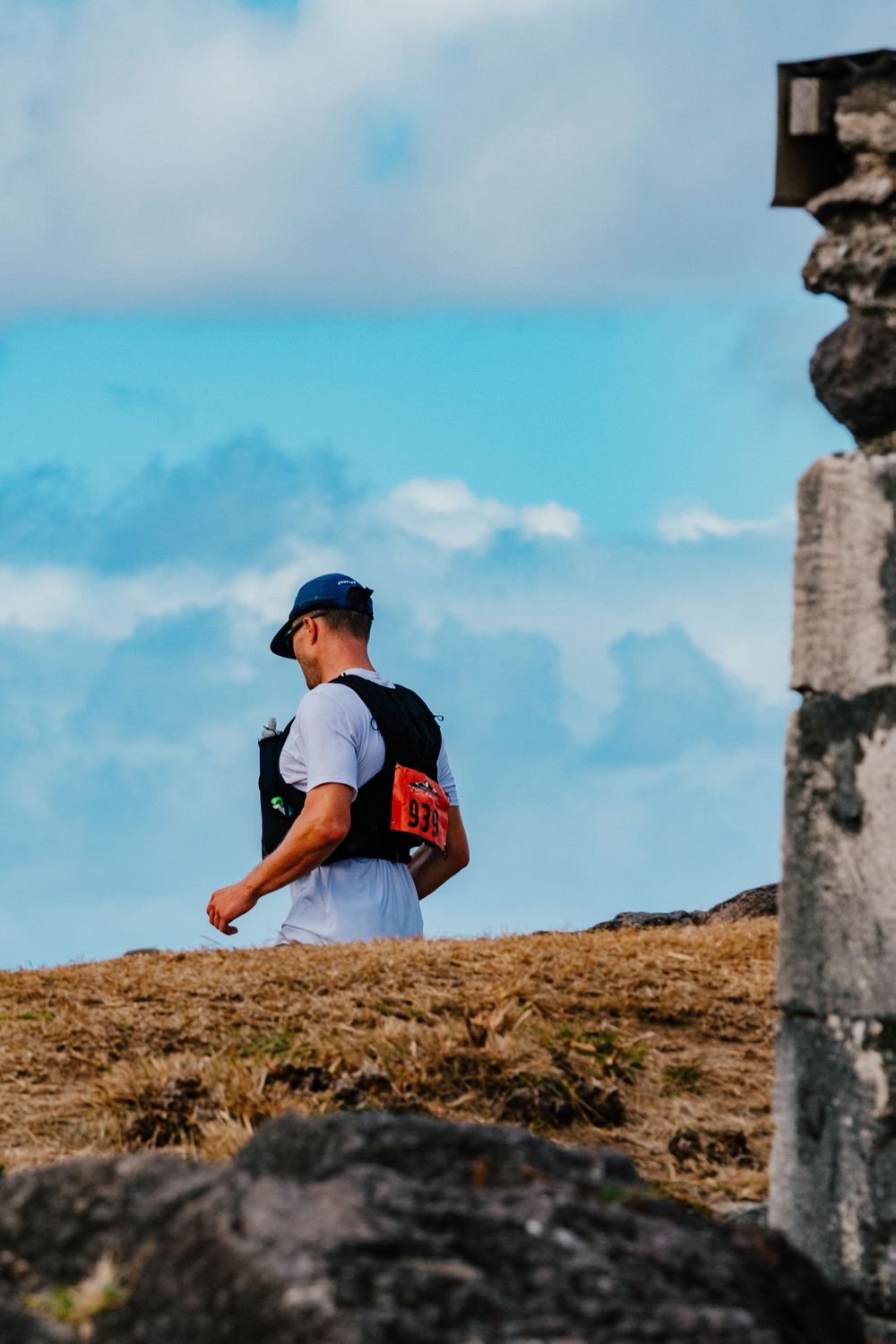 A hiker wearing a white shirt and cap stands on a rocky ridge against a bright blue sky with mountains in the distance.
