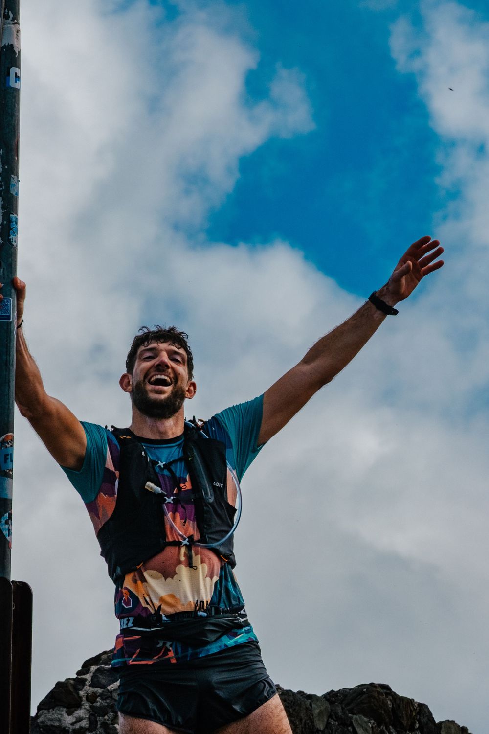 A person celebrating at the summit with outstretched arms against a blue cloudy sky background.