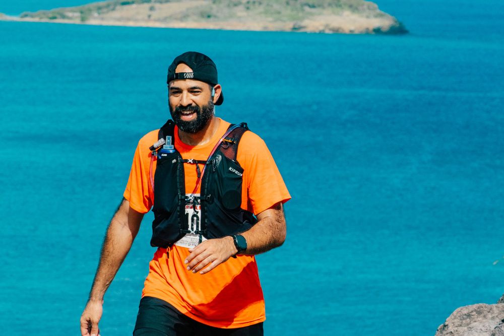 Athletic person in orange shirt and black life vest smiles while standing against turquoise ocean waters and rocky coastline.