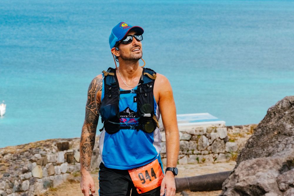 Athlete wearing blue tank top and hydration pack during trail run along coastal cliffs with ocean in background.