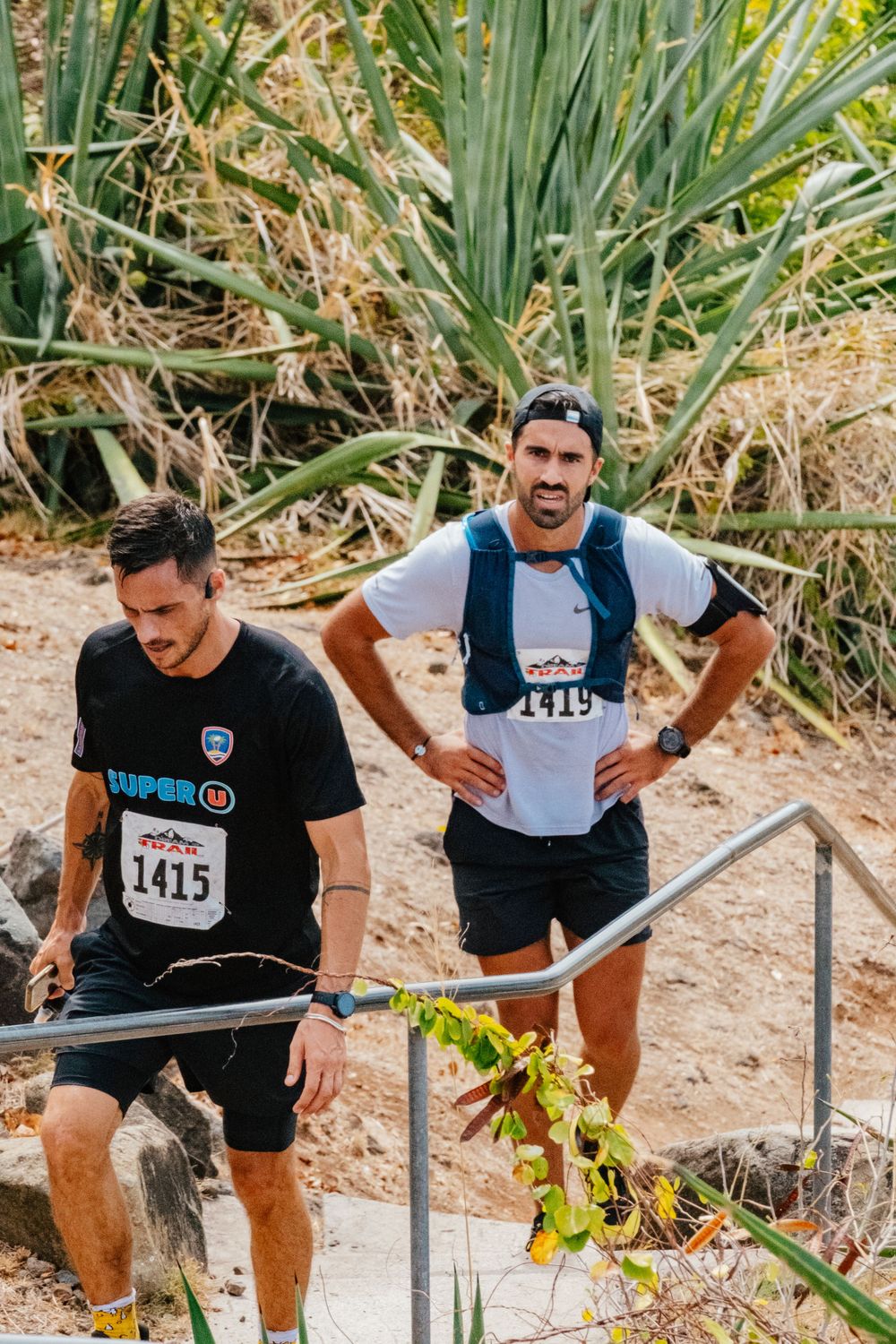 Two runners climb stone steps during an outdoor trail race with tropical plants in the background.