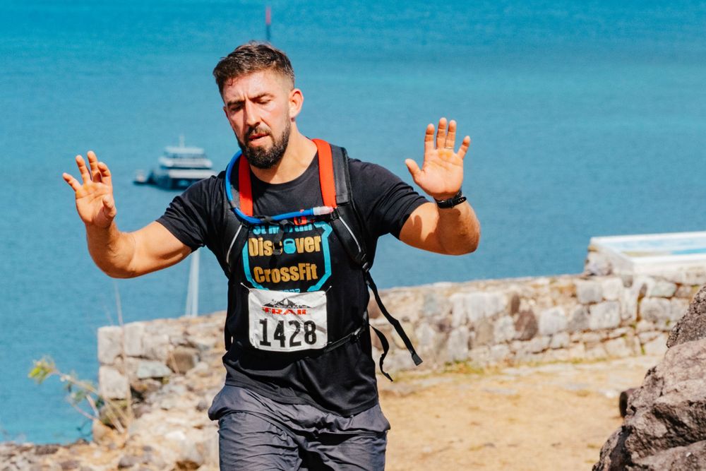 Runner with bib number 1429 gestures with arms raised along coastal trail with blue ocean and stone wall visible.
