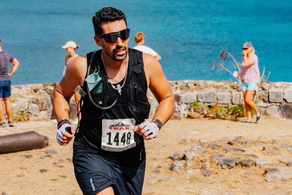Runner wearing sunglasses and black tank top participates in a beach race with blue ocean waters in background.