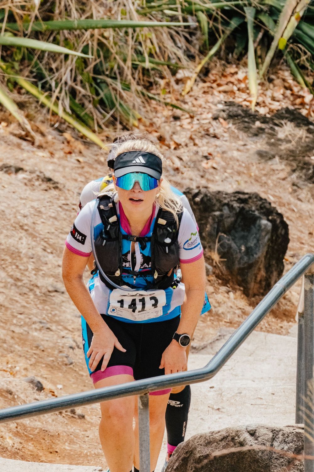 Athletic runner wearing sports gear and reflective sunglasses climbs uphill on rocky trail during endurance race.