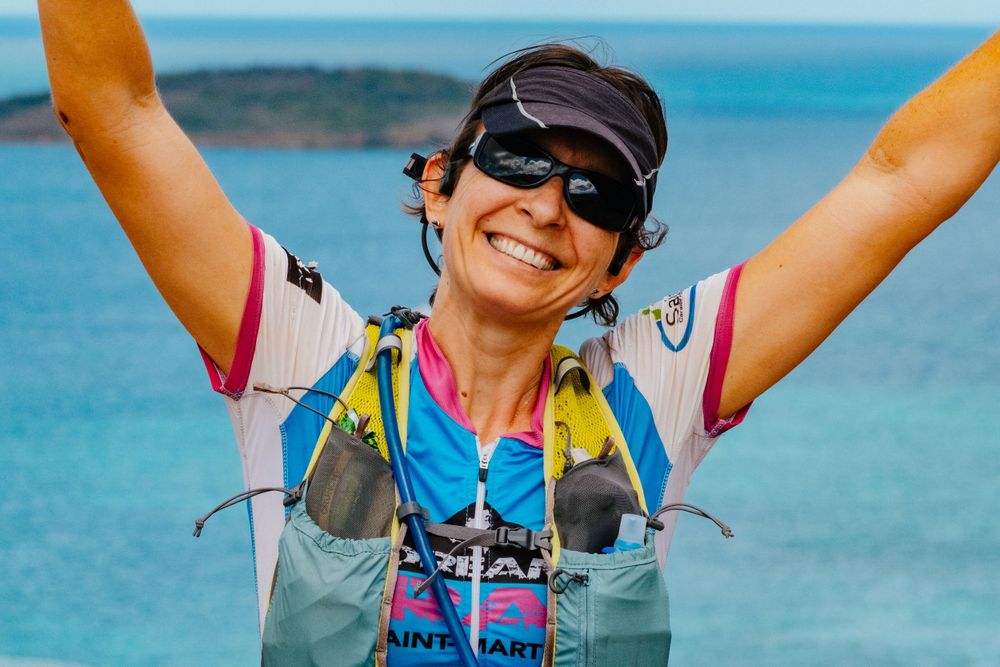 Joyful athlete wearing sunglasses and hydration vest celebrates with arms raised against bright blue ocean backdrop.
