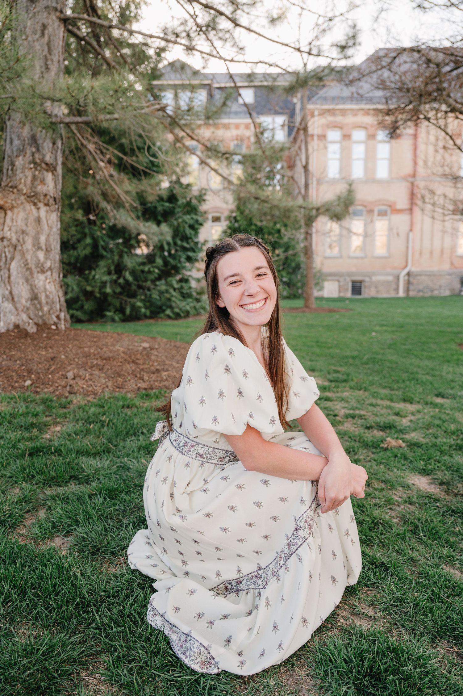 Someone in a white patterned dress sits on grass with a historic brick building visible in the background.