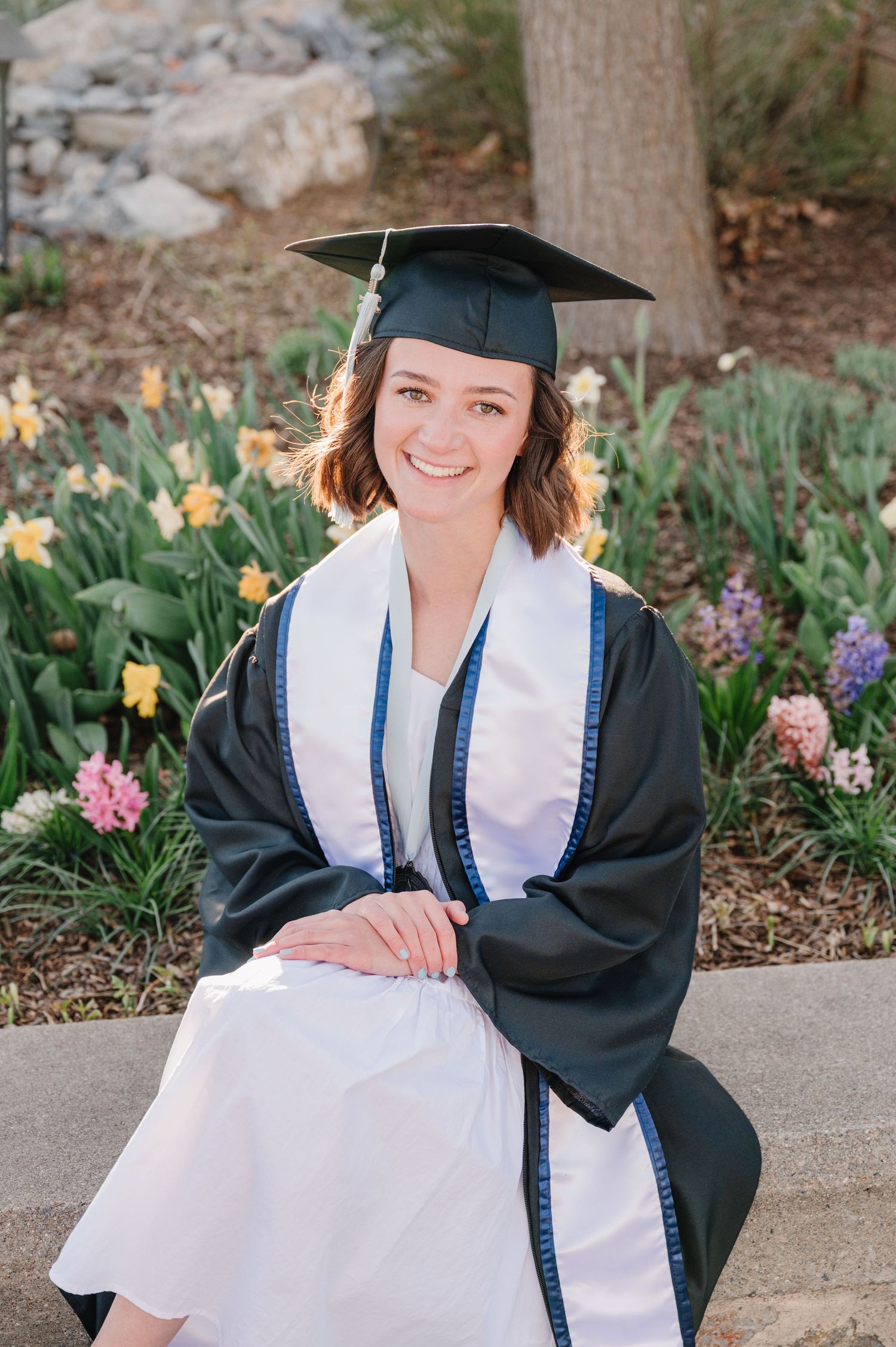 Graduate in cap and gown sits on concrete step surrounded by blooming daffodils and pink flowers in garden setting.