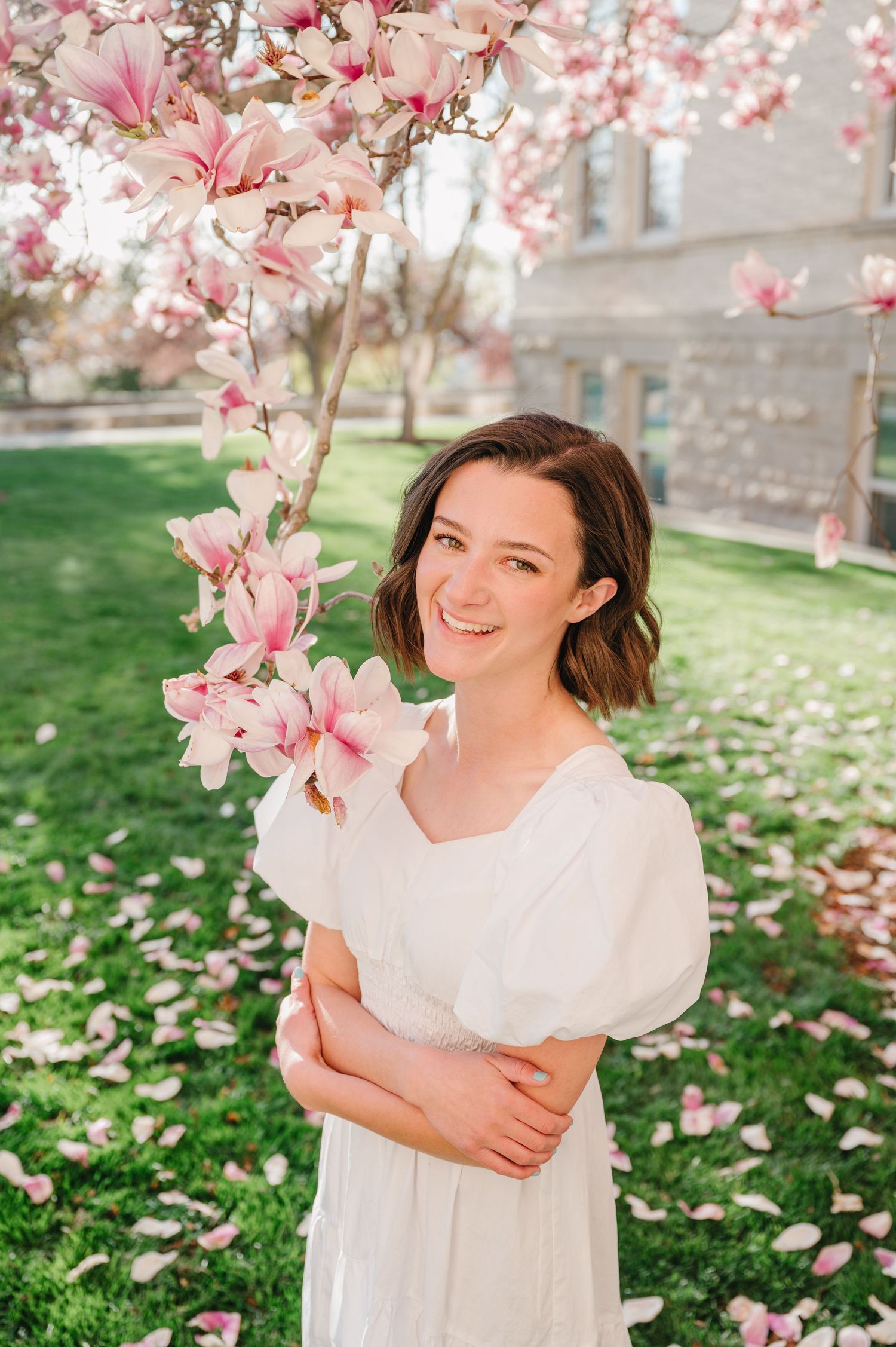 Beautiful spring scene with pink magnolia blossoms framing someone in a white dress on the grass by a stone wall.