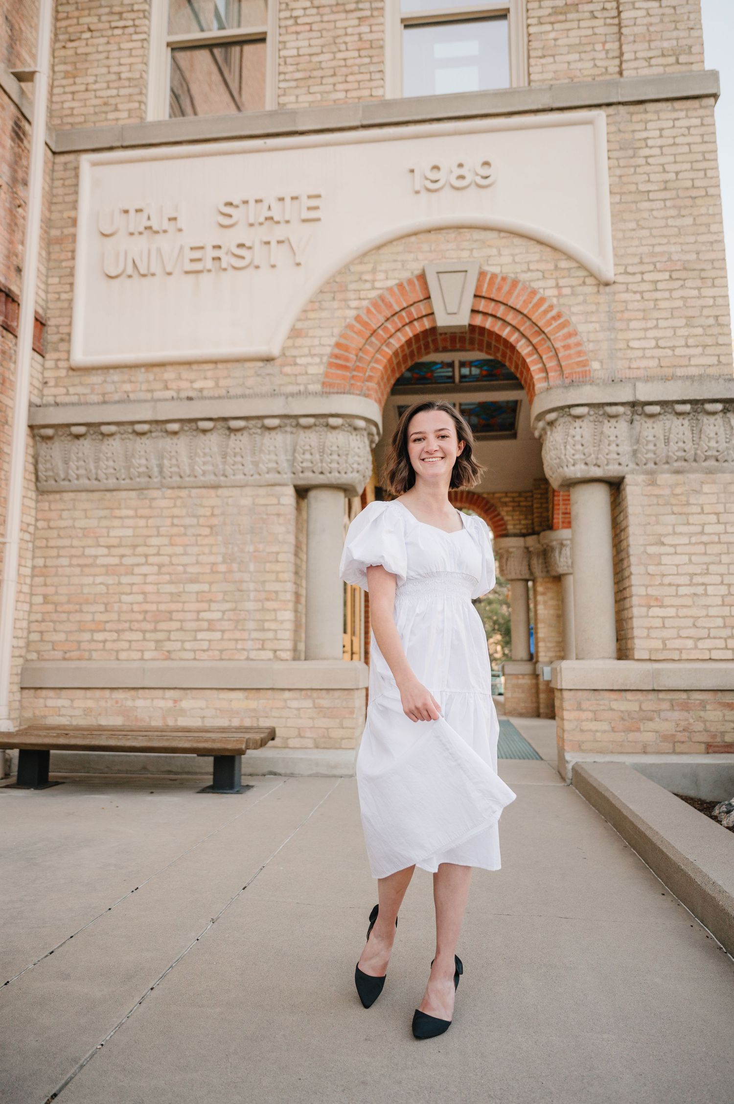 A white dress and black heels outfit captured in front of a vintage Utah State University building with brick facade.