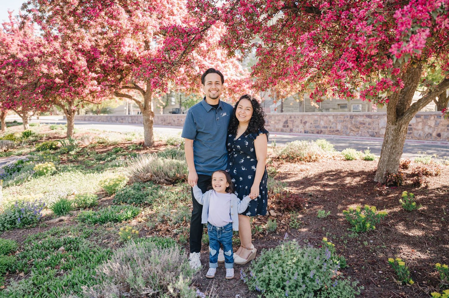 A family stands together under blooming pink cherry blossom trees on a sunny spring day in a garden setting.