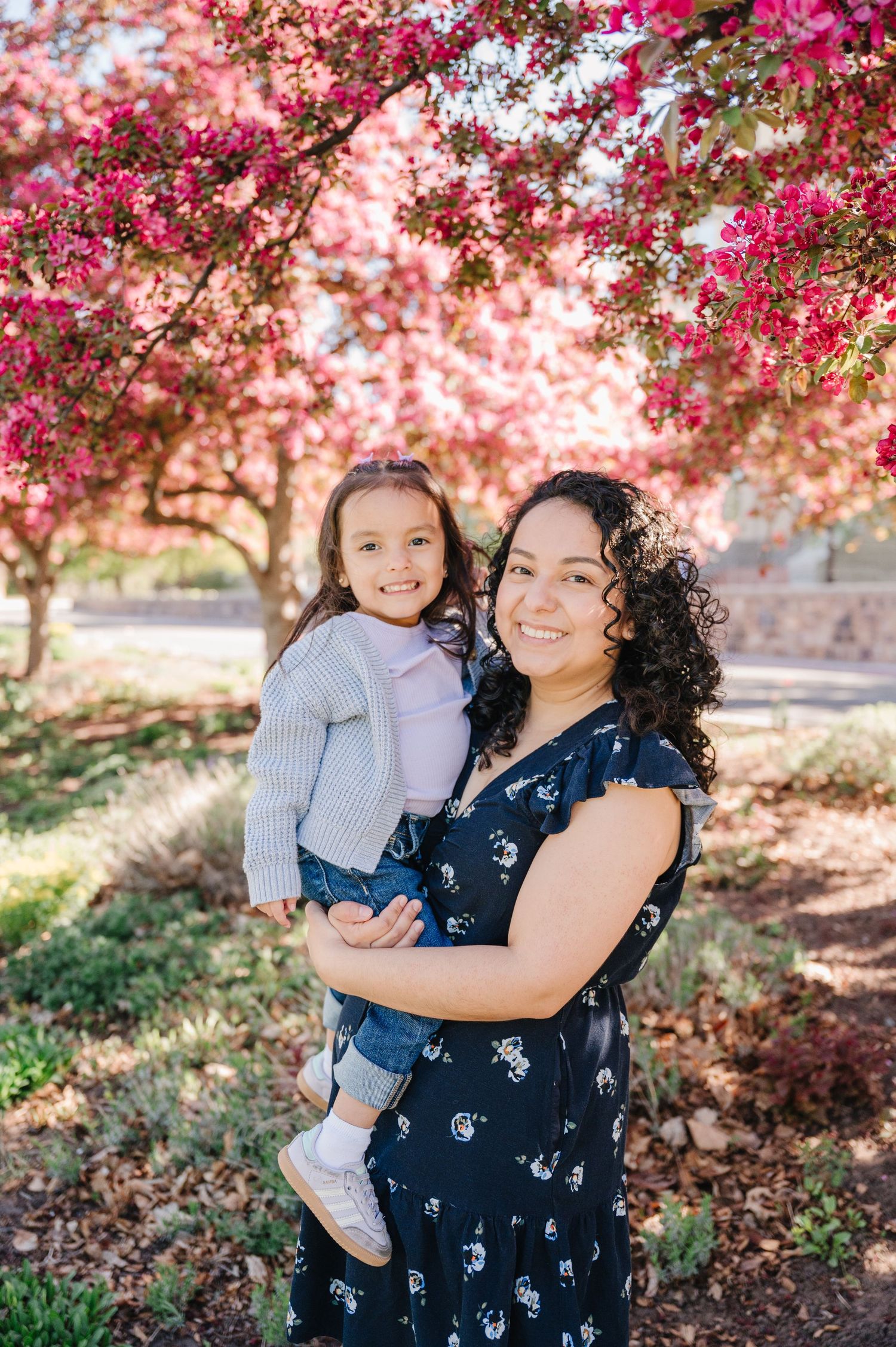 A mother and child pose together under blooming pink cherry blossom trees on a sunny spring day.