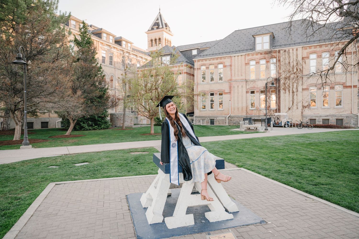 Graduate poses on architectural art installation on college campus with historic building in background.