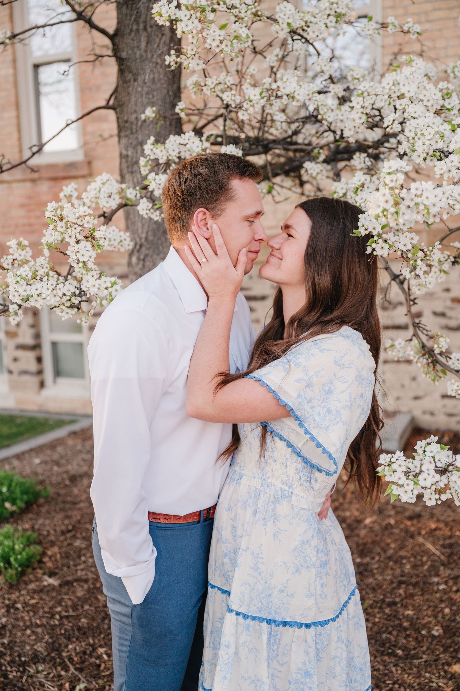 A romantic springtime moment under blooming white cherry blossoms against a rustic brick building wall.