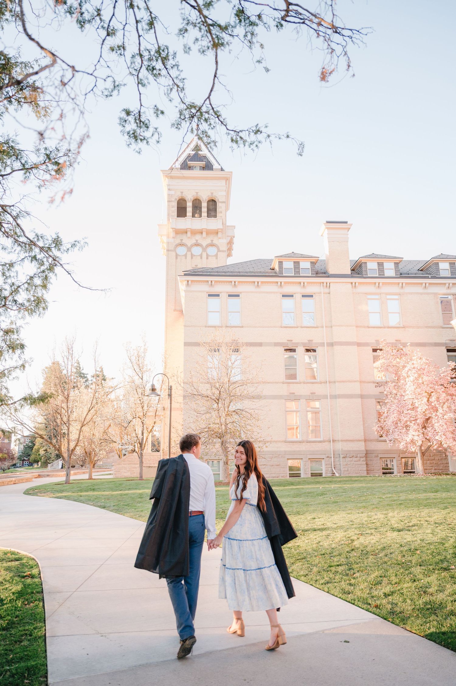 A couple walks hand in hand along a pathway by a historic university building with a tower on a sunny autumn day.