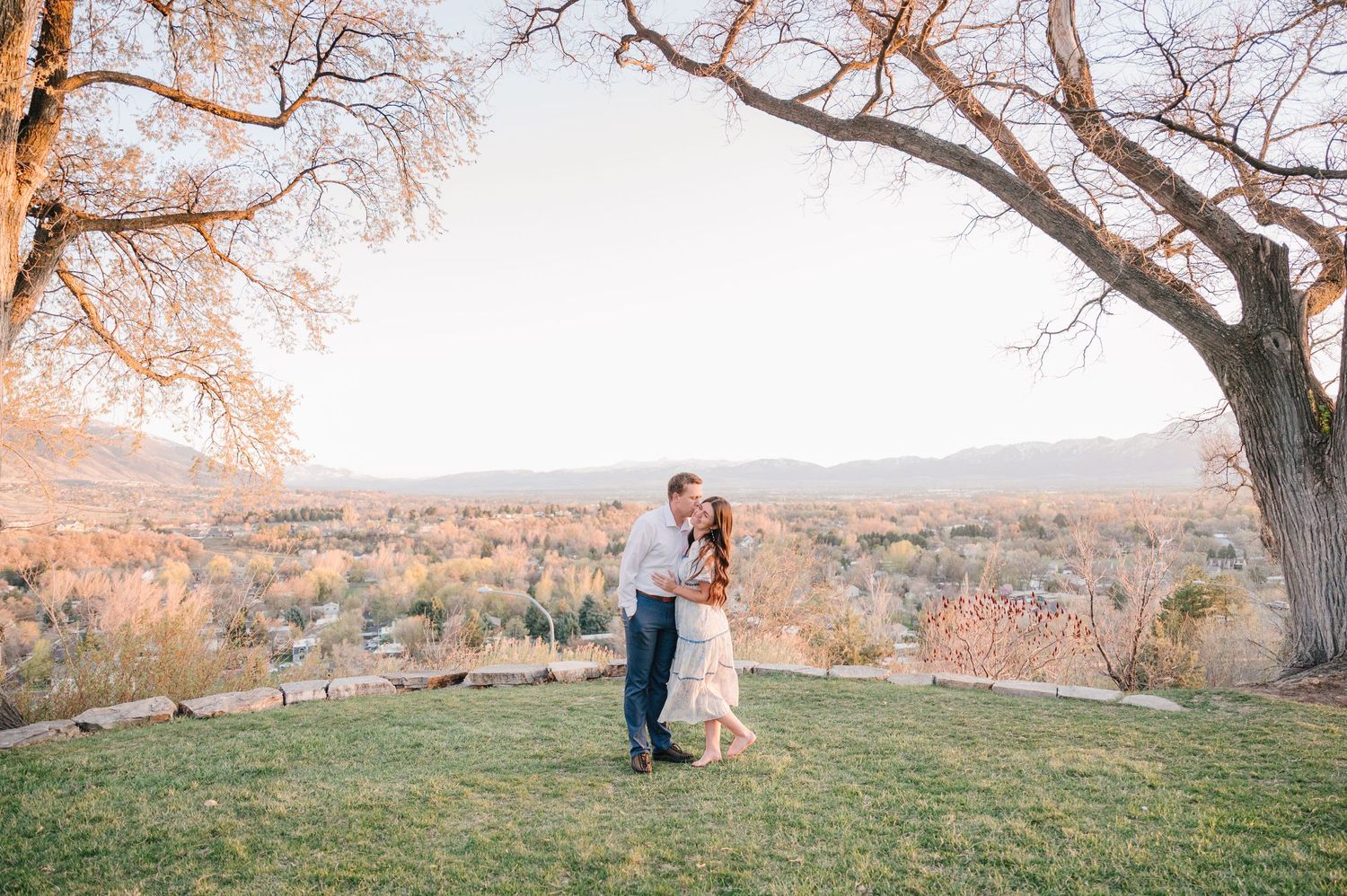 A couple shares a romantic moment on a hilltop overlooking an autumn landscape at sunset under a large tree.