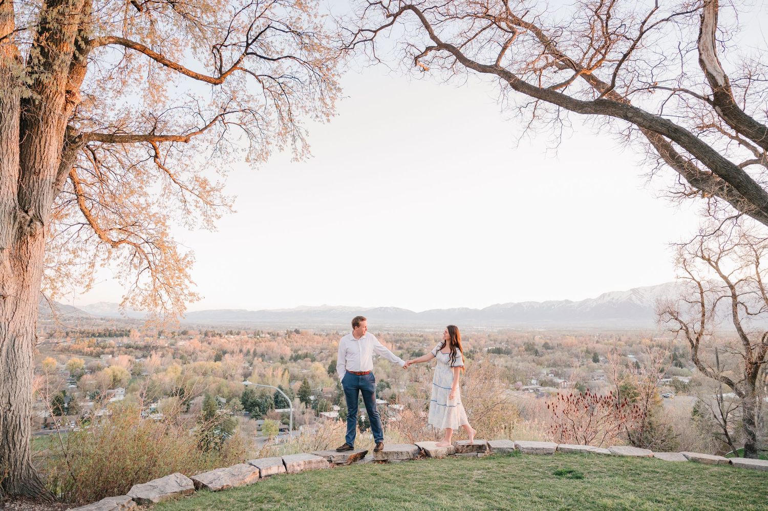 A couple stands on a hilltop overlooking a scenic valley at sunset, framed by bare tree branches above.